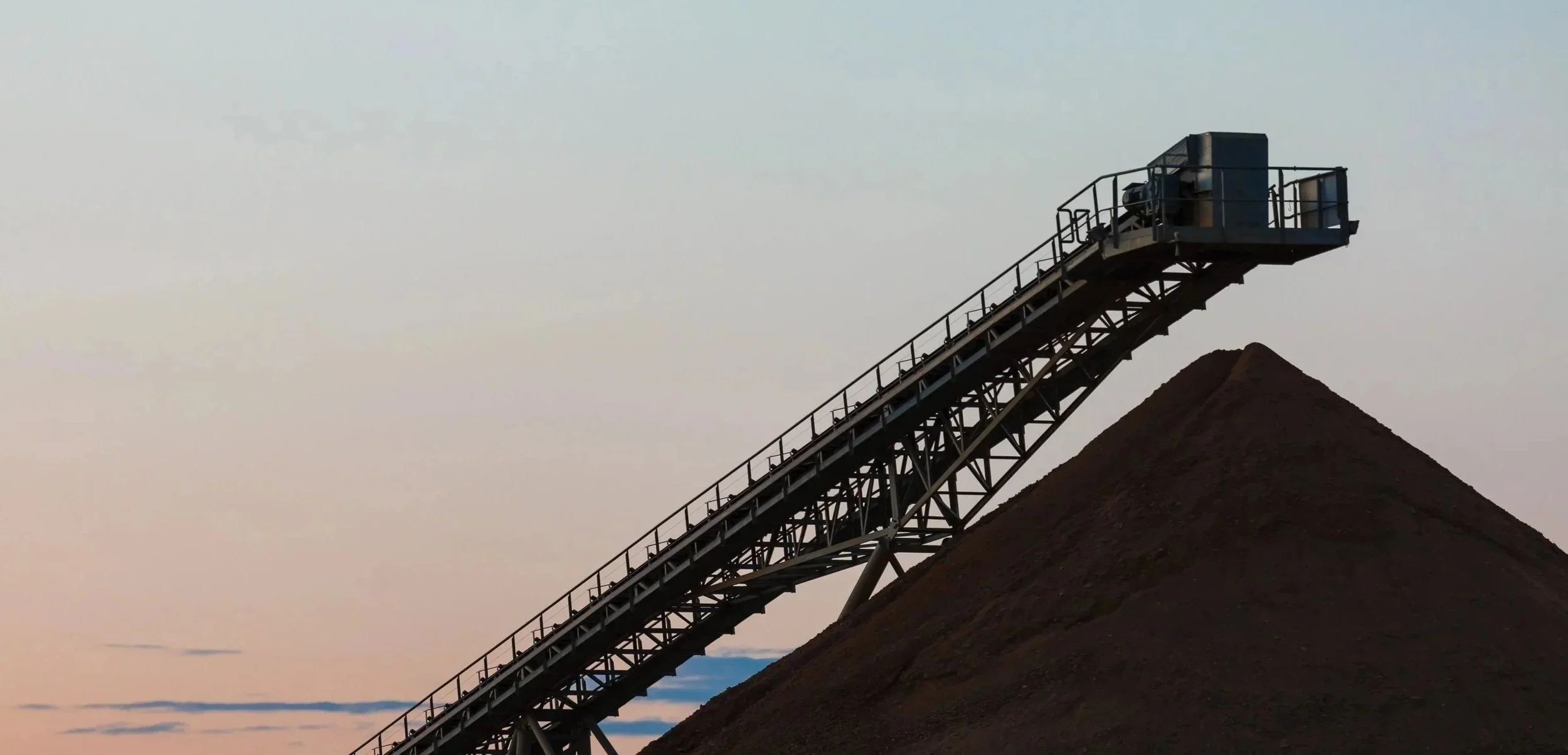 A conveyor belt extending upward over a mound of dark earth at sunset.