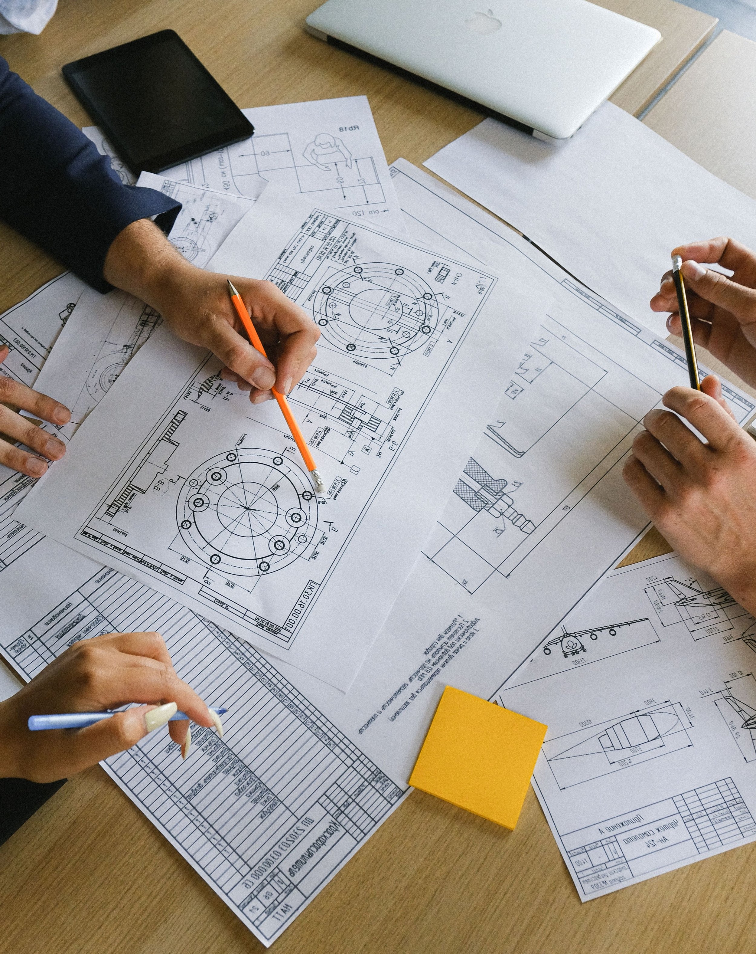 Several technical drawings and blueprints for mechanical parts are scattered on a wooden table, with some hands pointing at different sections, and a closed laptop and notebooks visible in the background.