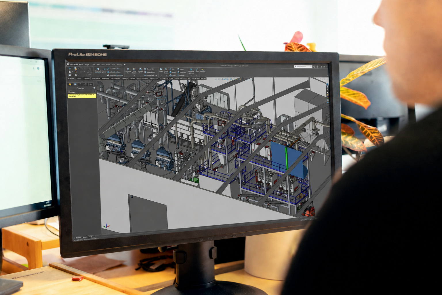 A person looking at a computer screen displaying a 3D engineering design of an industrial facility with pipes and machinery.