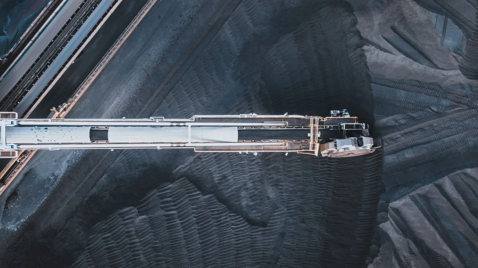 An aerial view of a large industrial conveyor system moving dark mineral or coal across a mining site with visible tire tracks and excavated land.
