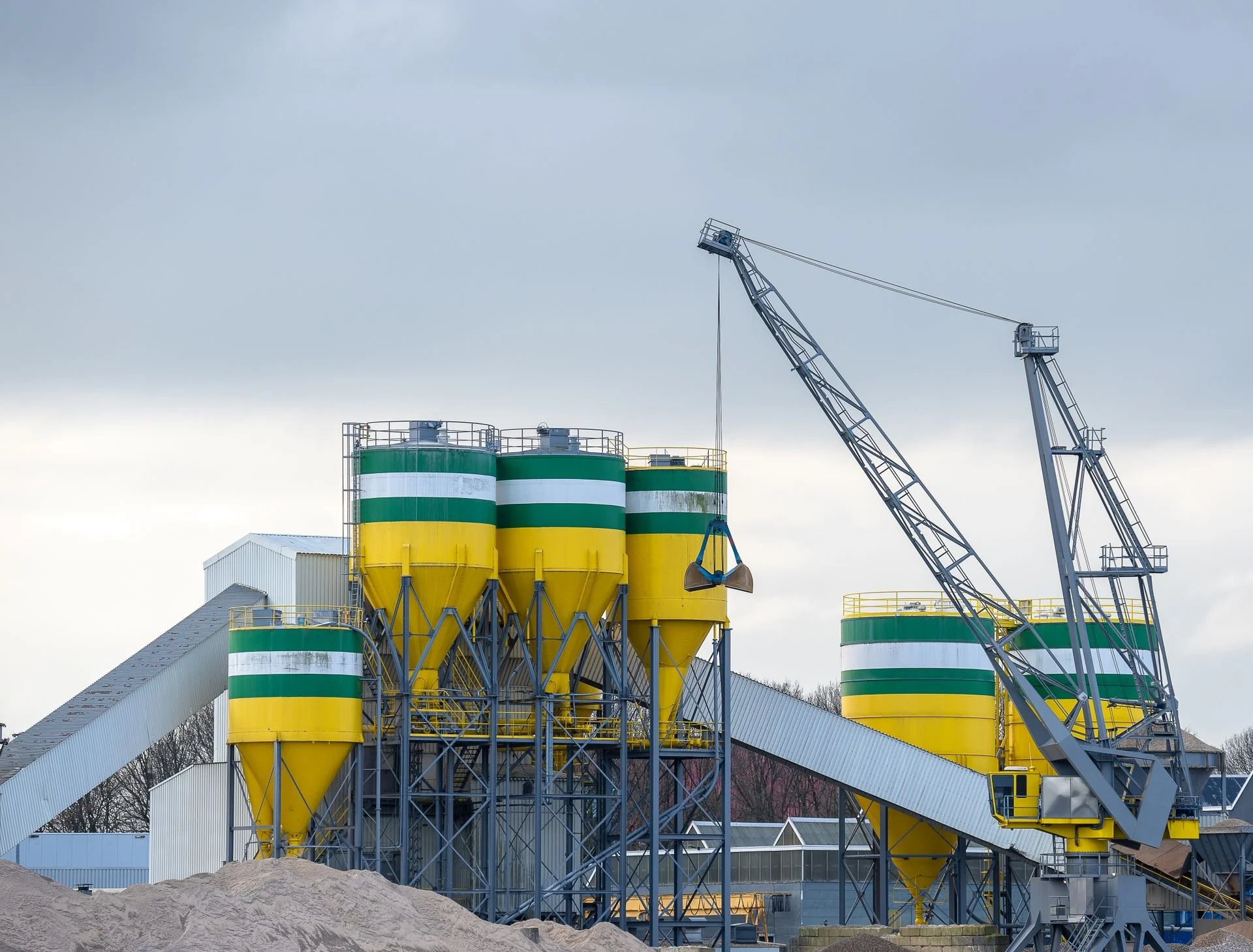 Industrial facility with yellow, green, and white storage silos, a large conveyor system, and cranes against a cloudy sky.