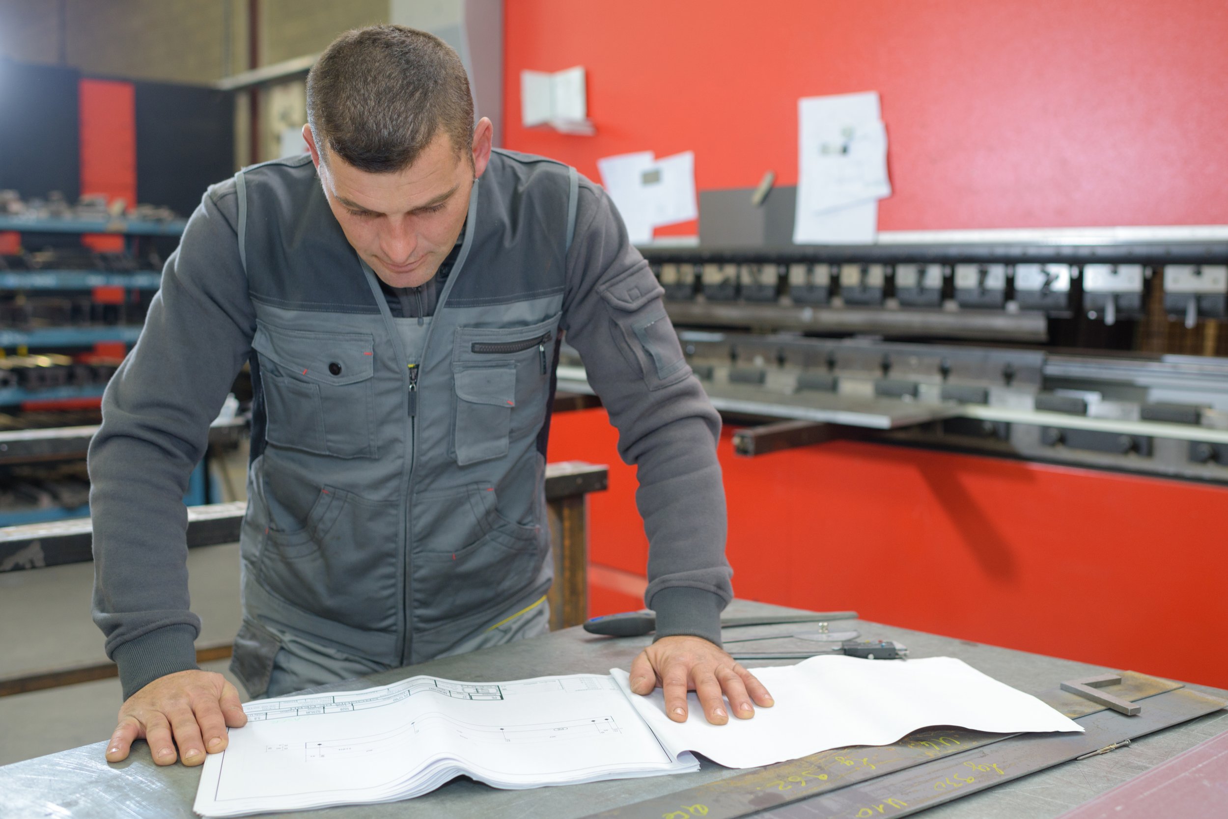 A man in a gray uniform looking down at architectural blueprints on a table in a workshop with red and gray walls and tools around him.