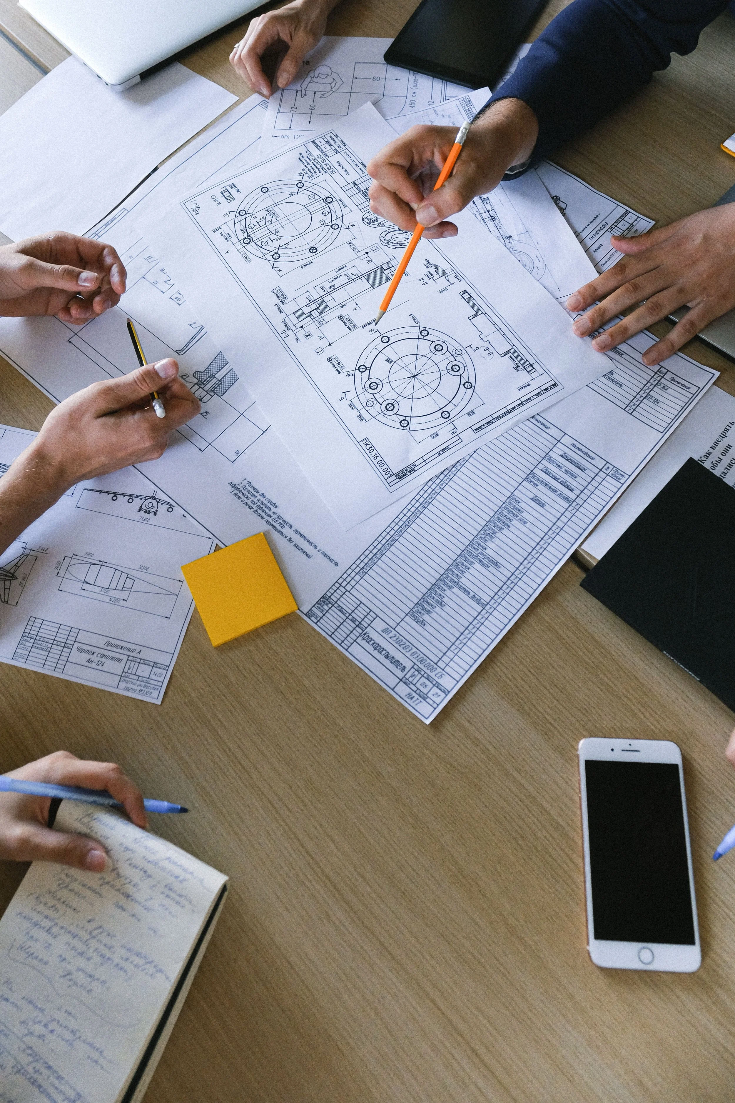 People working on technical drawing blueprints, discussing engineering or mechanical design plans on a wooden table, with notebooks, a smartphone, and various documents scattered around.