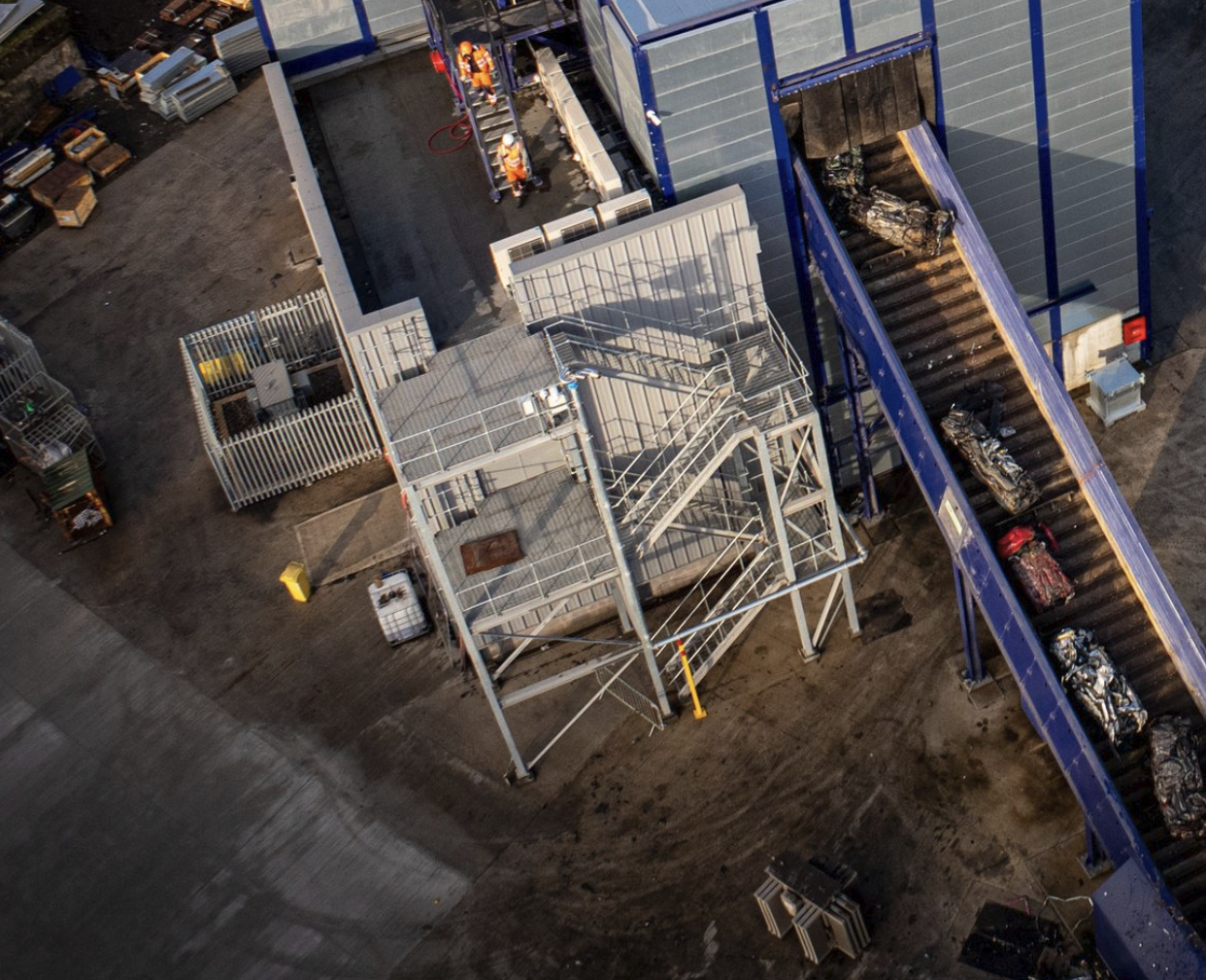Overhead view of an industrial outdoor area showing a baggage claim conveyor belt with wrapped luggage, a metal staircase, workers in safety vests, and various storage or work areas.