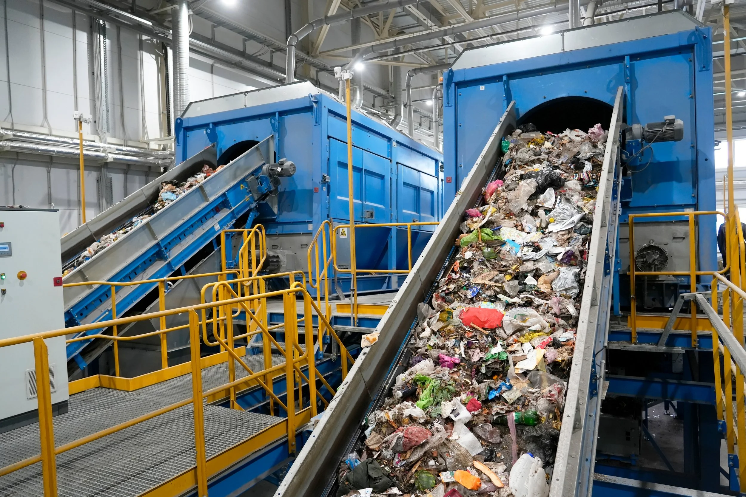 Industrial recycling facility with blue trommel screen machines and conveyor belts carrying municipal waste and recyclables.