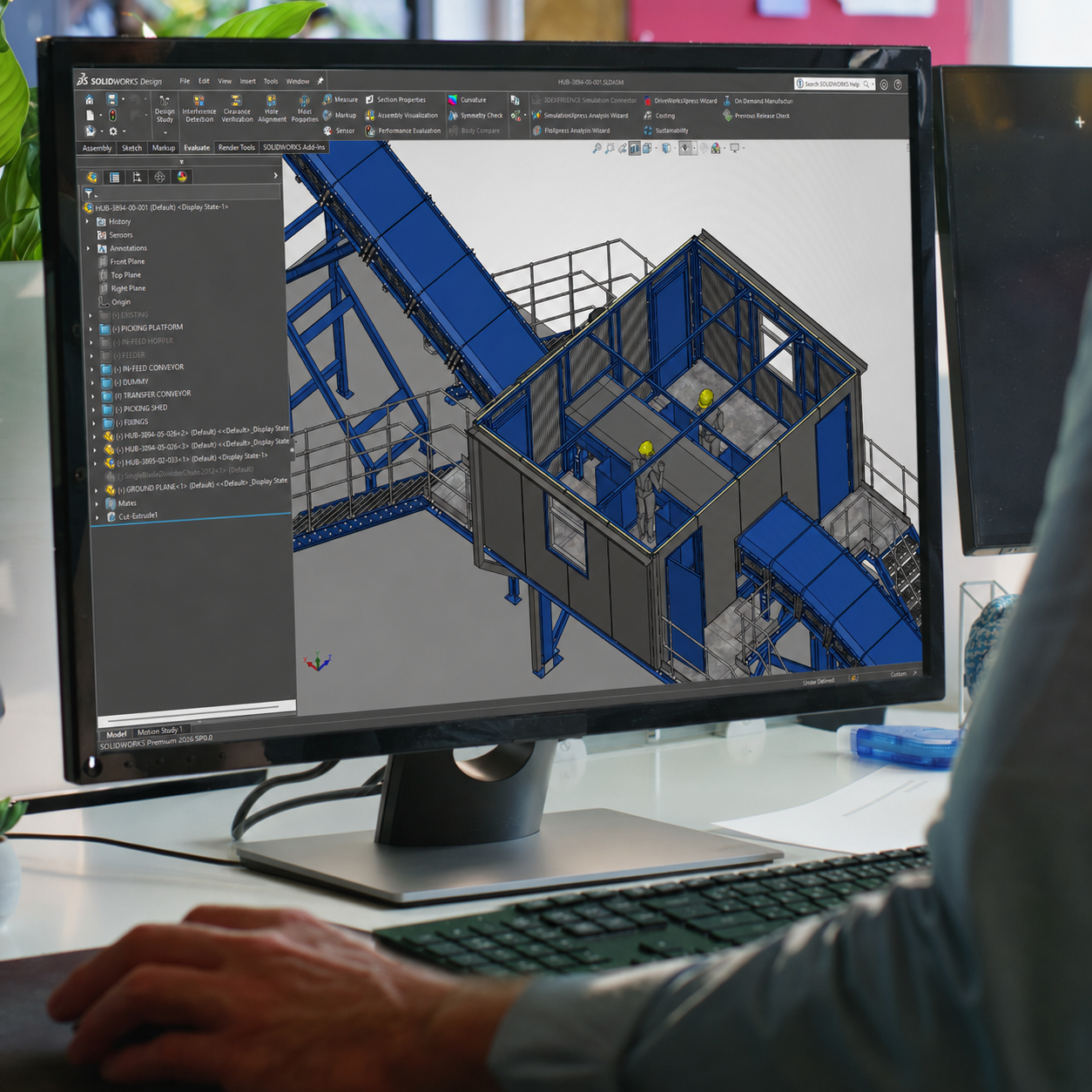 Computer screen displaying a 3D CAD model of an industrial automated conveyor system with a blue framework and safety rails. Person working at a desk with a keyboard, mouse, and a pen visible.