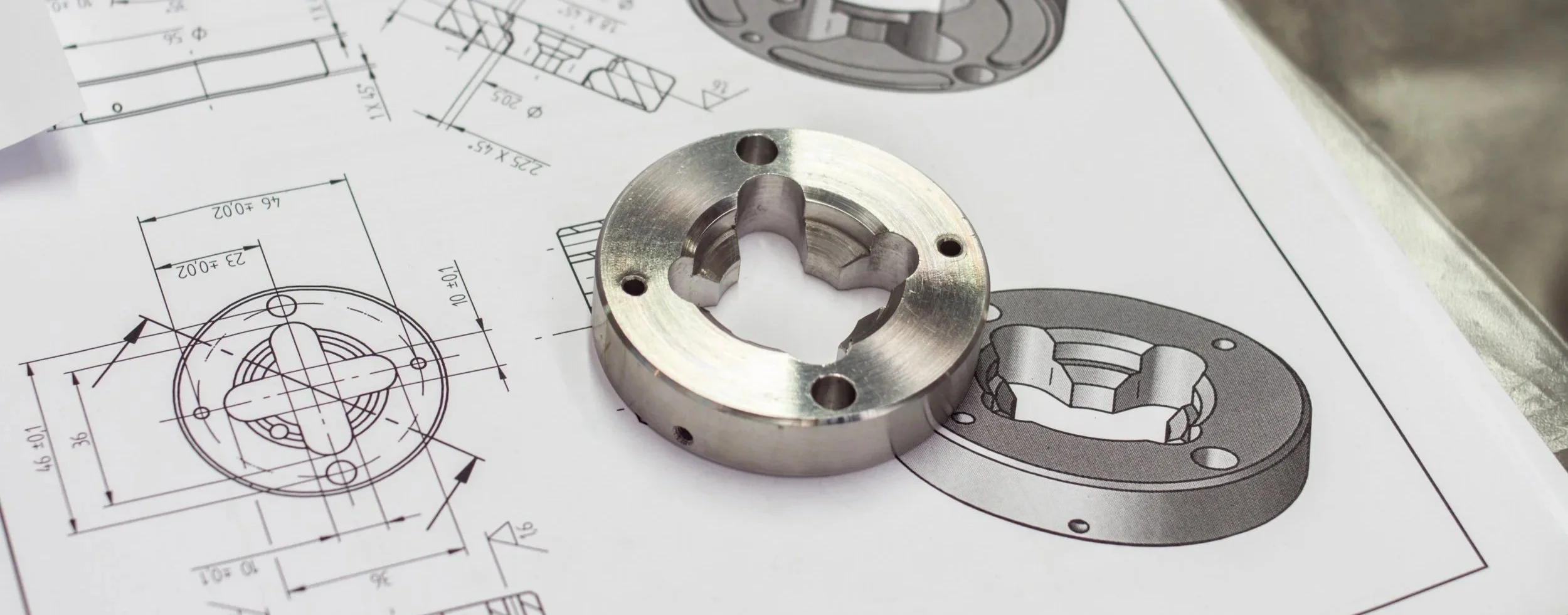 Metal mechanical part placed on technical engineering drawings with detailed diagrams and measurements.