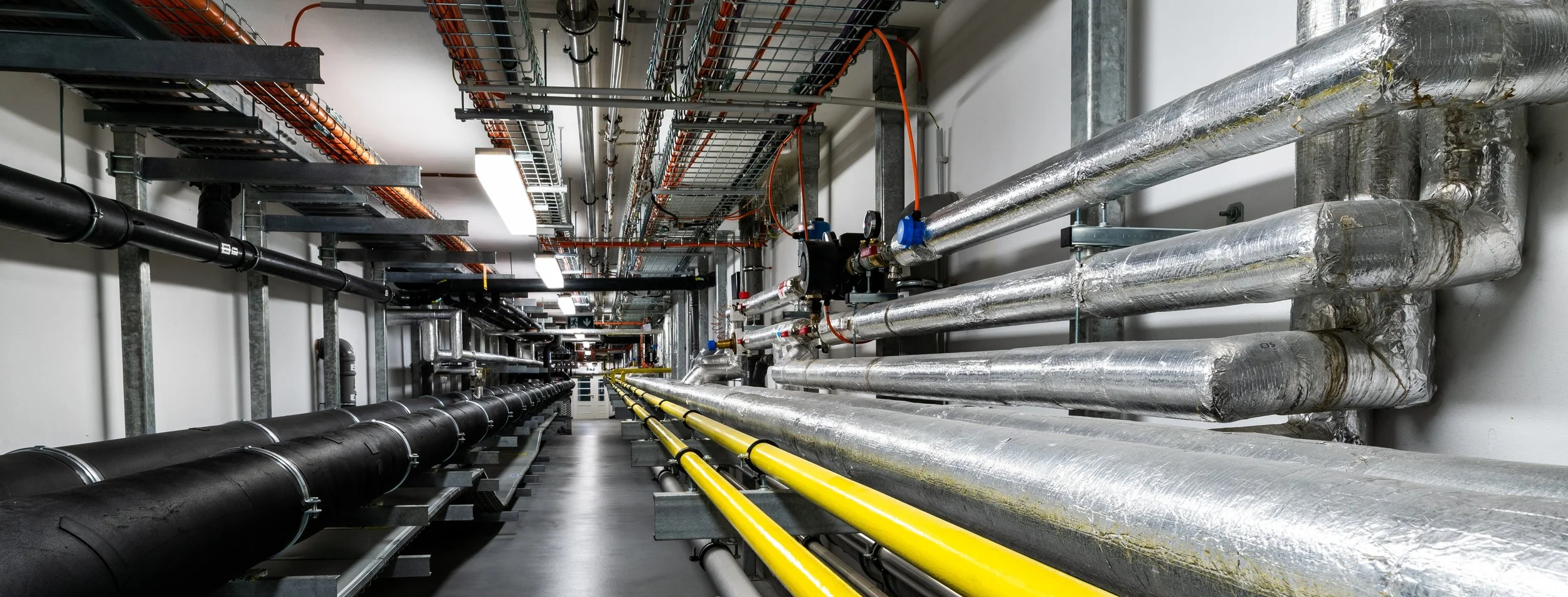 A facility room with industrial pipes, black and yellow pipelines, metal conduits, and wiring on the ceiling, used for plumbing or electrical systems.