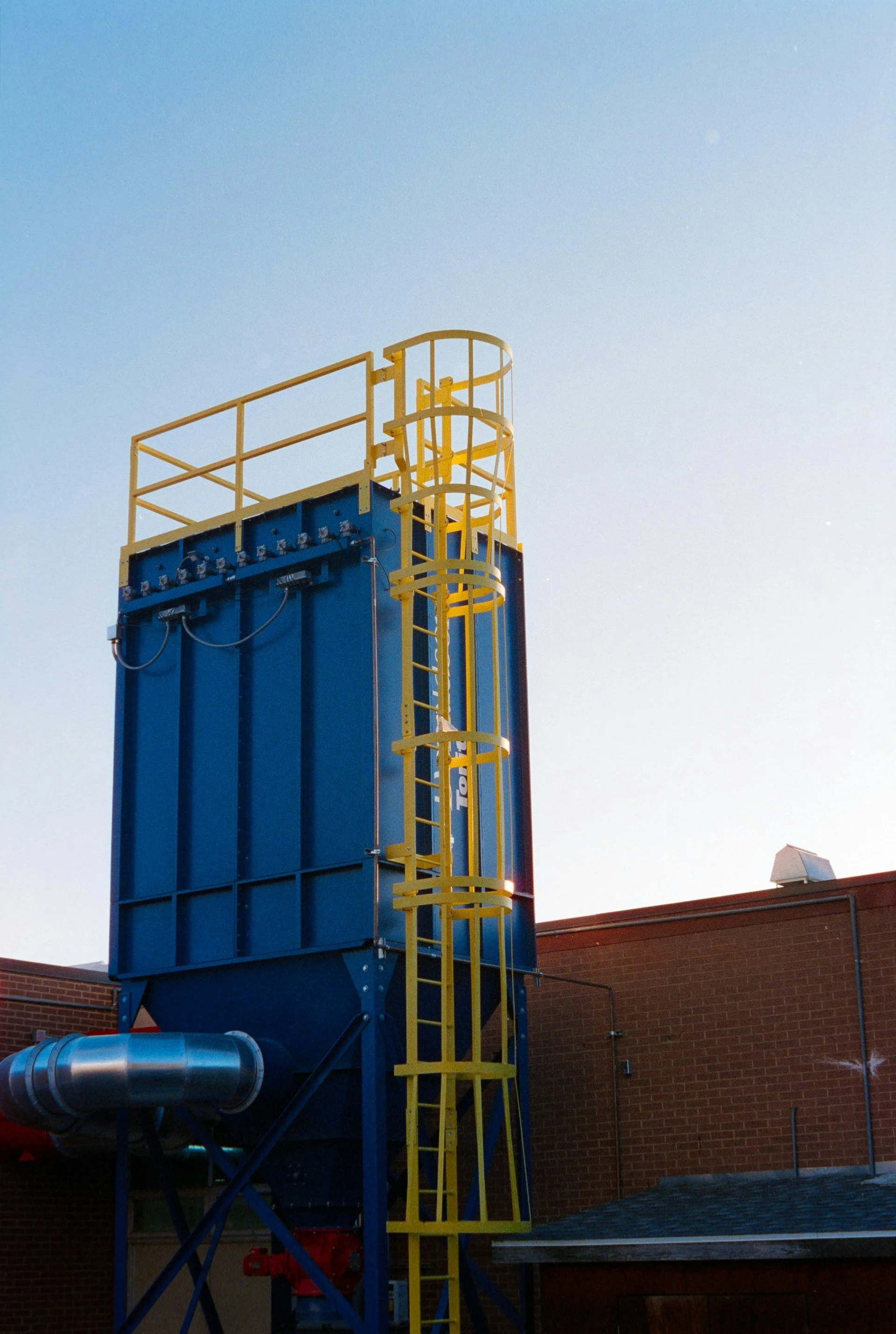 A tall blue and yellow hydraulic lift structure with a safety railing at the top, located outdoors against a brick building under a clear sky.