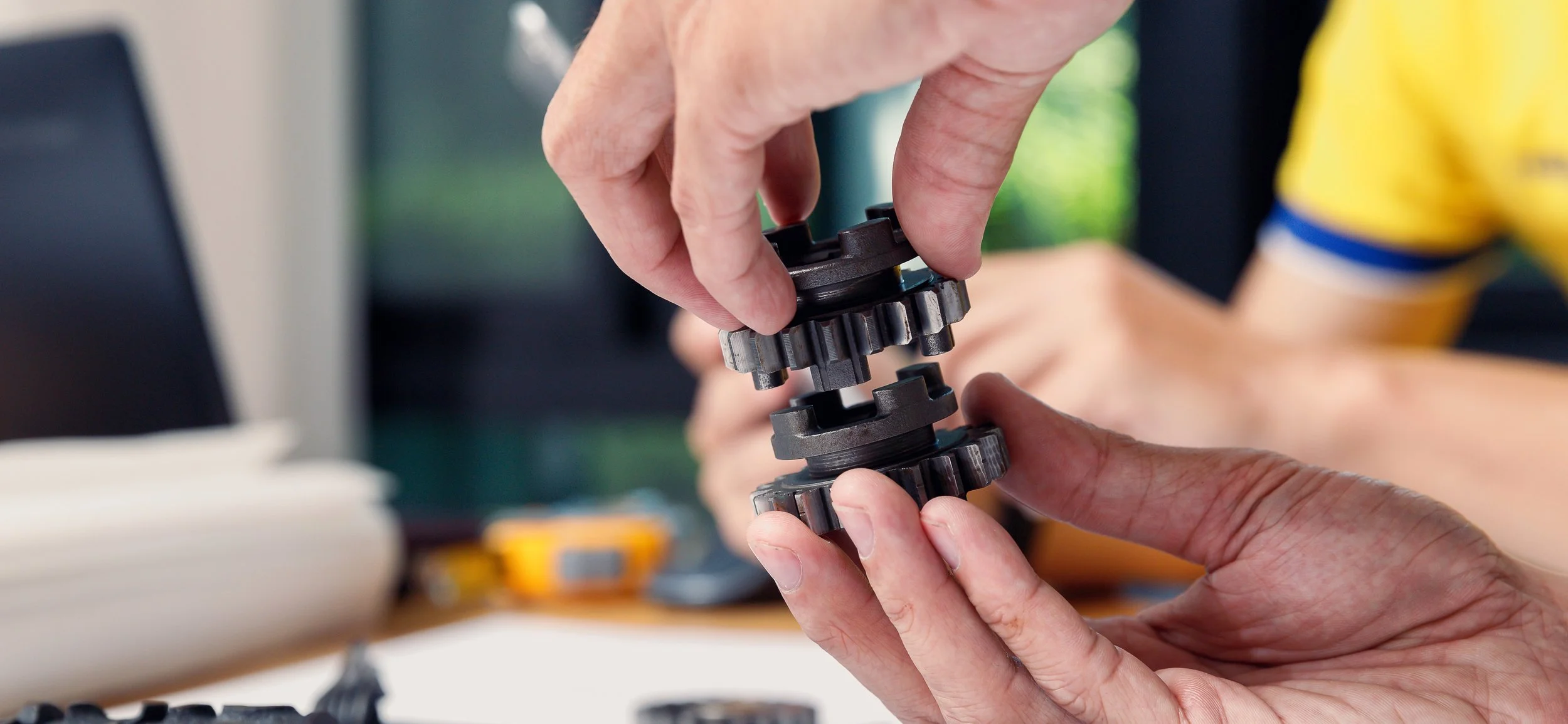 Close-up of hands assembling gears or mechanical parts, with a blurred background showing a workspace.