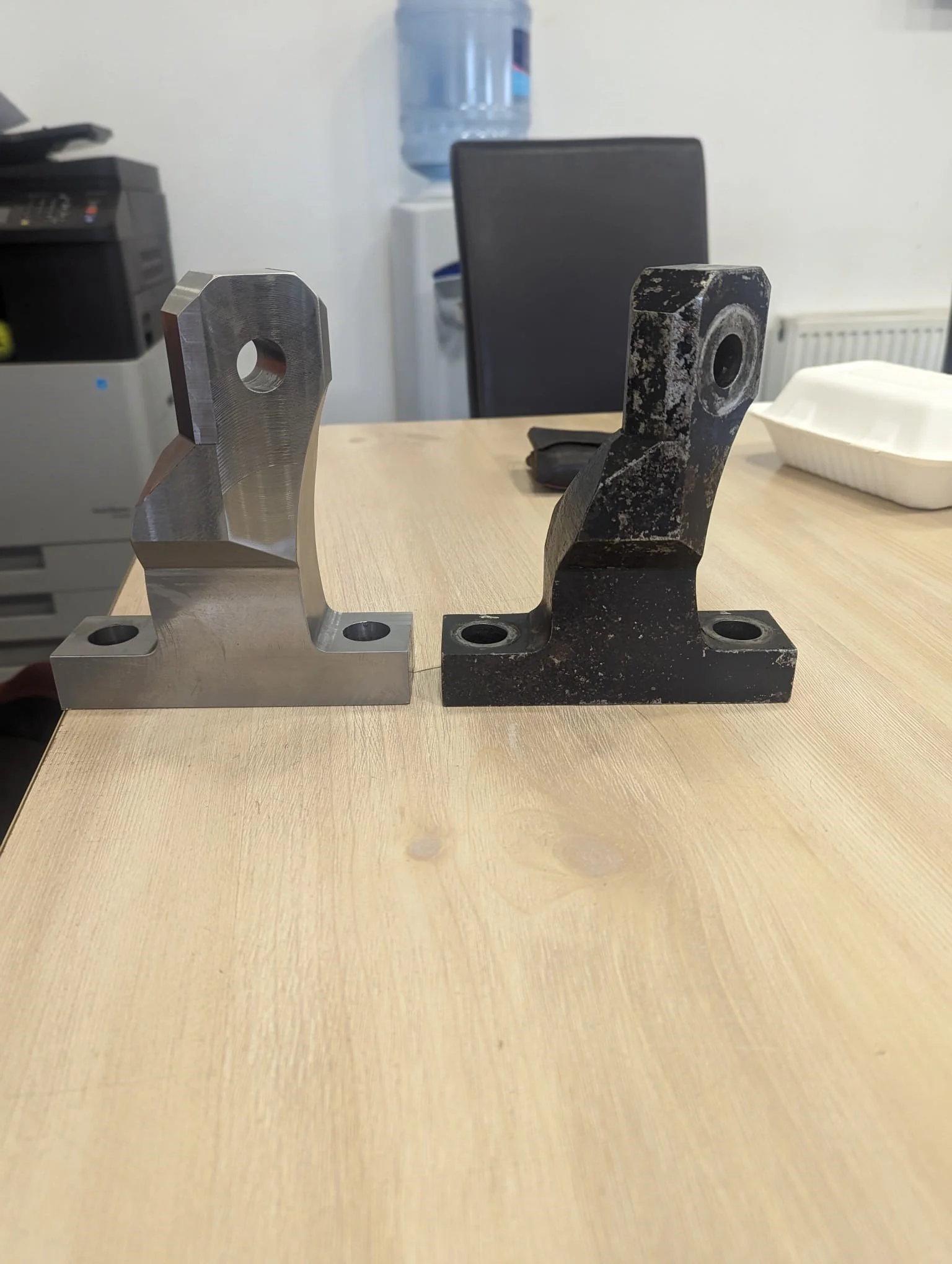 Two metal brackets placed on a wooden table with a water dispenser and office chair in the background.