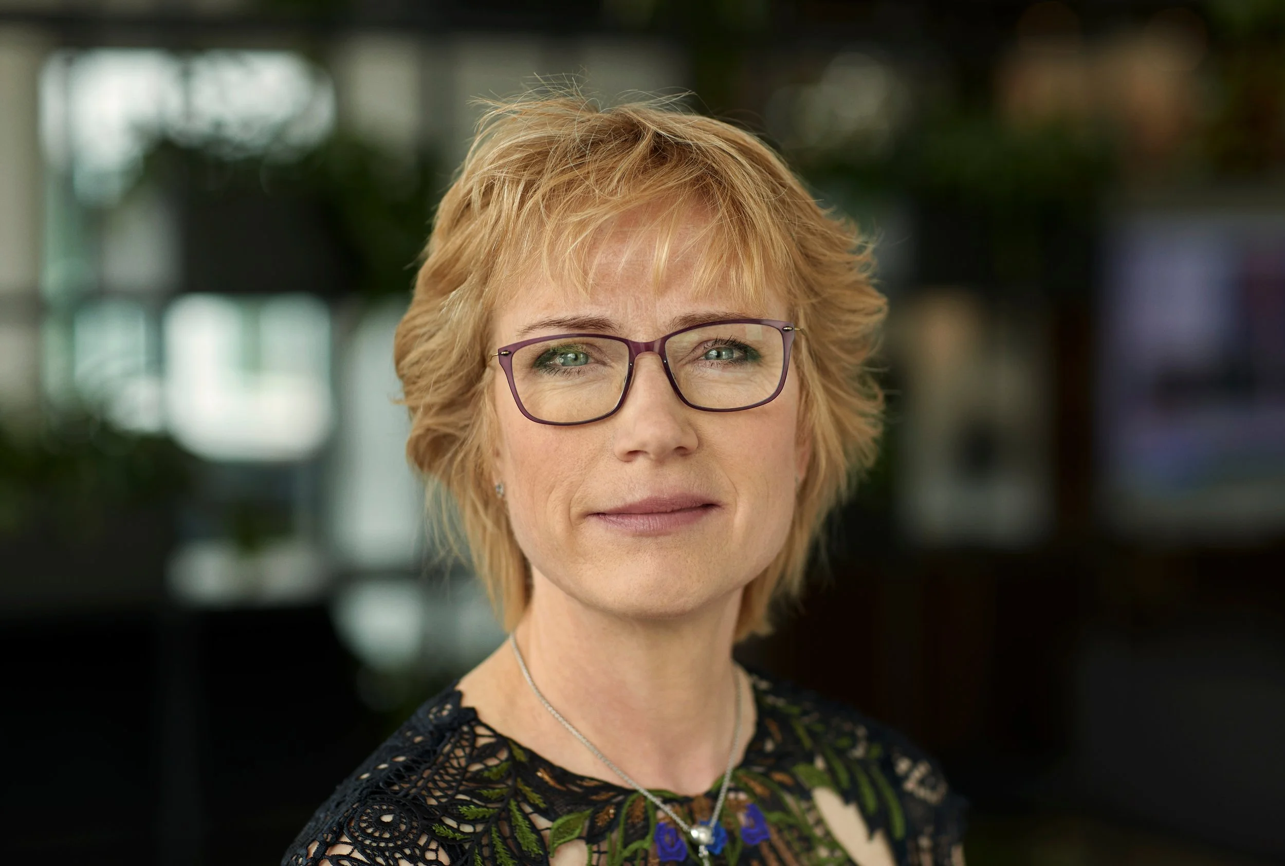 Et portræt af en  kvinde med blond, kort, bølget hår og briller, iført en sort kjole med broderi. Baggrunden er sløret og museal, med bløde lysreflekser. Dorte Krak, CEO hos Arp-Hansen Hotel Group