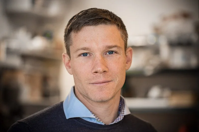 Portræt af en mand med kort brunt hår og blå øjne, iført en lys blå skjorte og en sort sweater, i et professionelt miljø. Christian Arp-Hansen, Projektleder for Orø Naturarv.