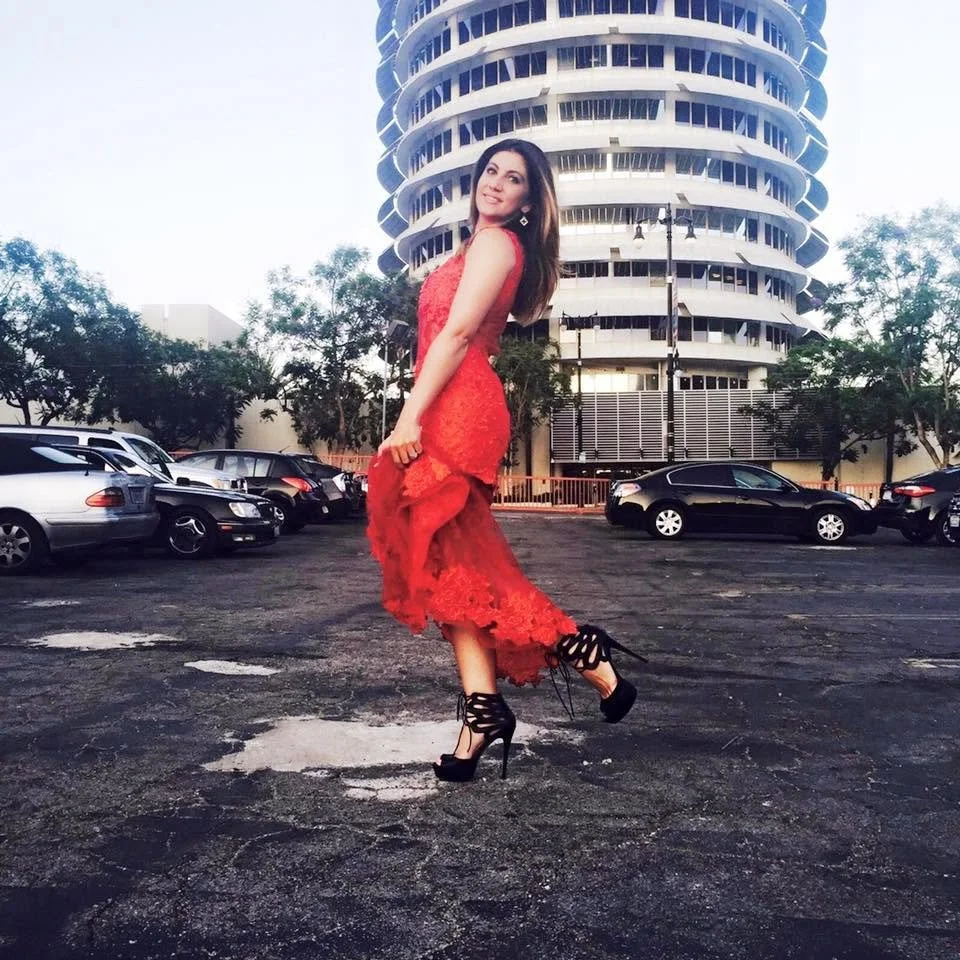 A woman in a red lace dress and black high heels poses in a parking lot with a modern, circular building in the background.