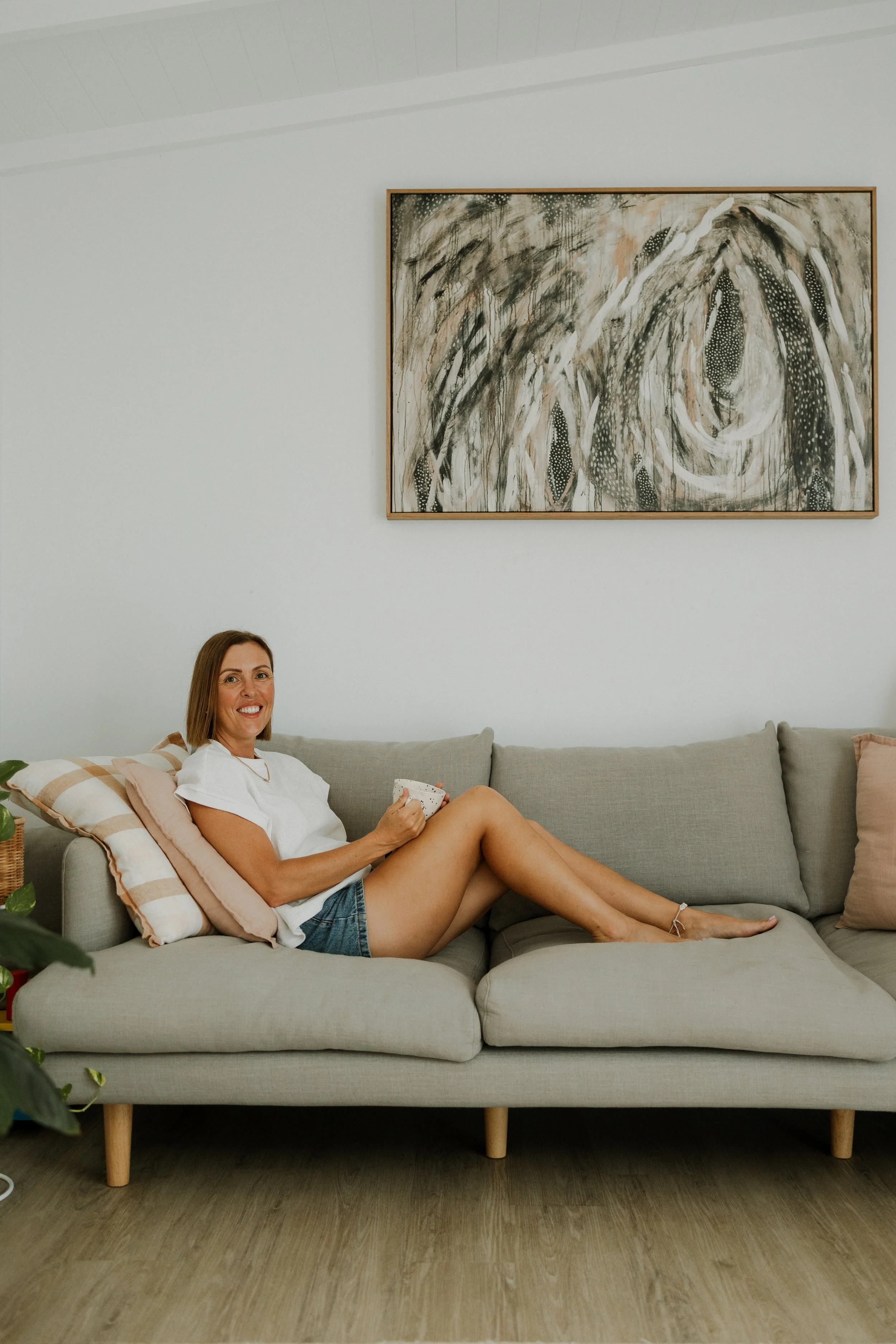 A woman with brown hair and a white shirt is sitting on a light gray sofa with beige and pink cushions, holding a bowl. She is smiling and wearing denim shorts, with her legs crossed. The living room has a wooden floor, and there is a large abstract painting with black, white, and brown tones hanging on the white wall behind her. Part of a green potted plant is visible in the bottom left corner of the image.