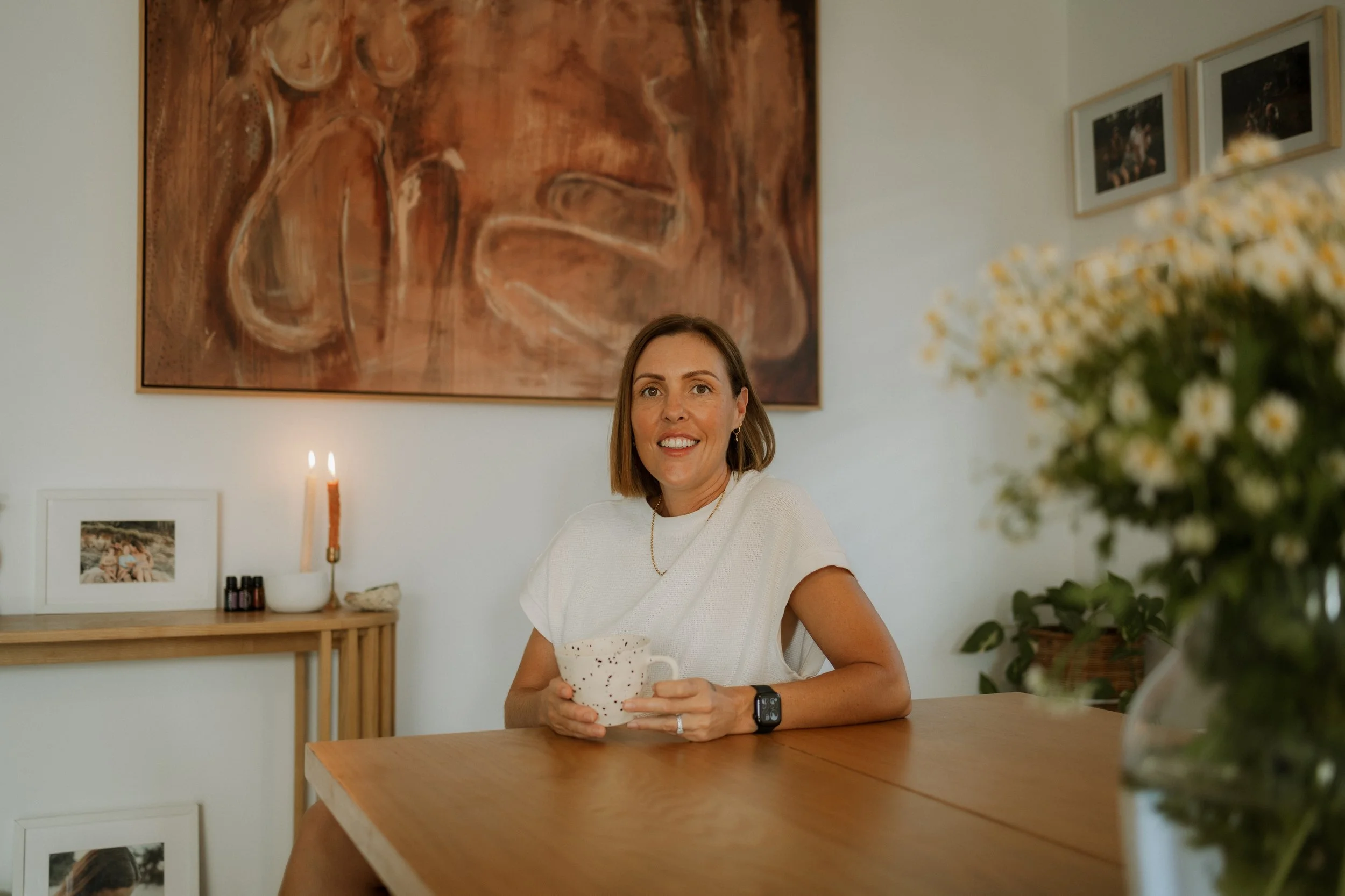 A woman sitting at a wooden table, holding a white mug with black speckles, smiling at the camera. She is wearing a white top, a watch on her left wrist, and earrings. In the background, there's a wall with a large abstract painting, framed photographs, a candle, and small bottles on a shelf. A plant with white flowers is partially visible on the right.