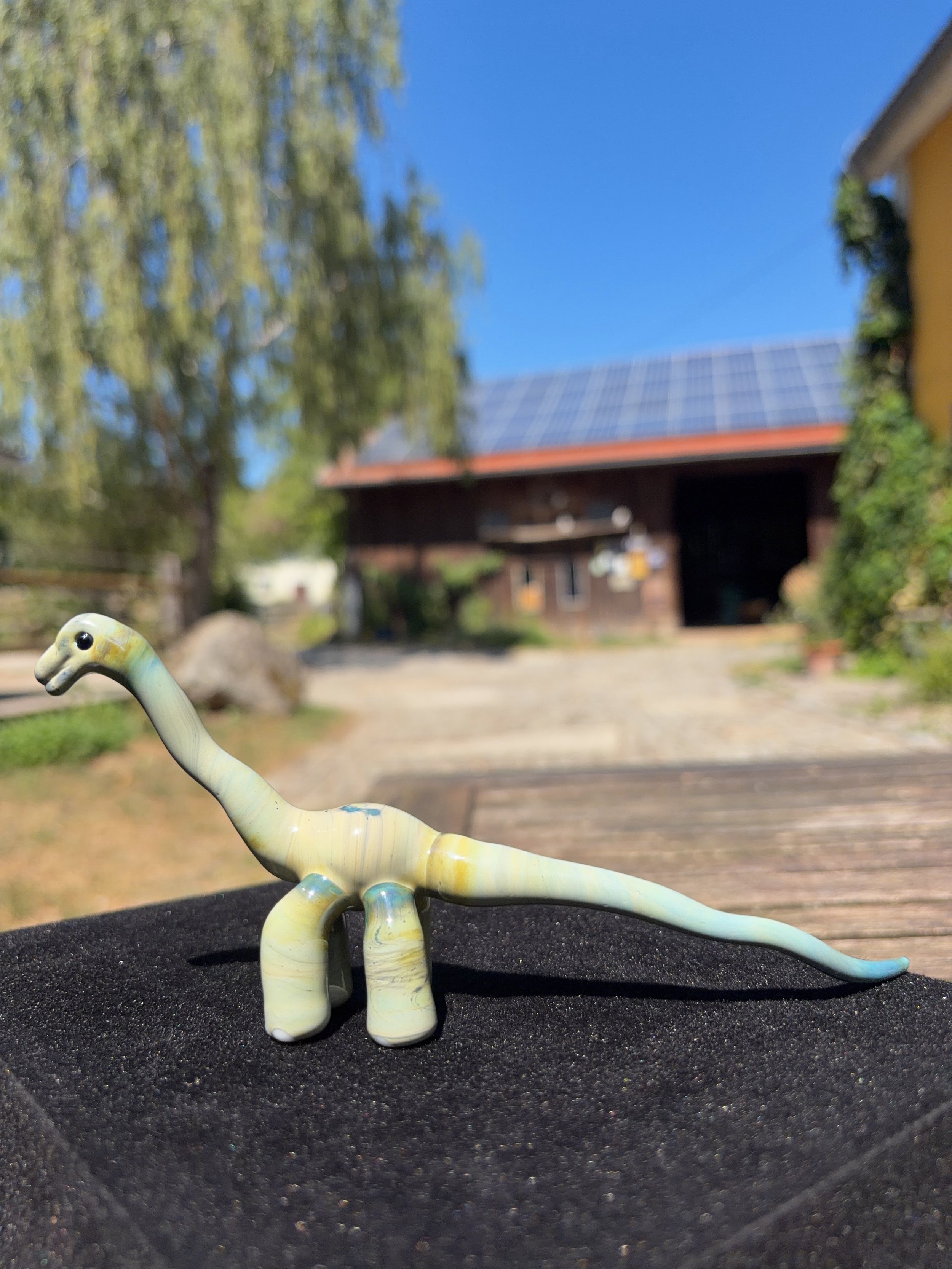 Miniatur-Brachiosaurus-Skulptur aus Glas vor Landschaft, StudioJoshiKunstfigur eines grünen, langhalsigen Dinosauriers auf schwarzem Untergrund, im Hintergrund ein Bauernhaus mit Solarpanelen auf dem Dach, Baum und blauer Himmel.