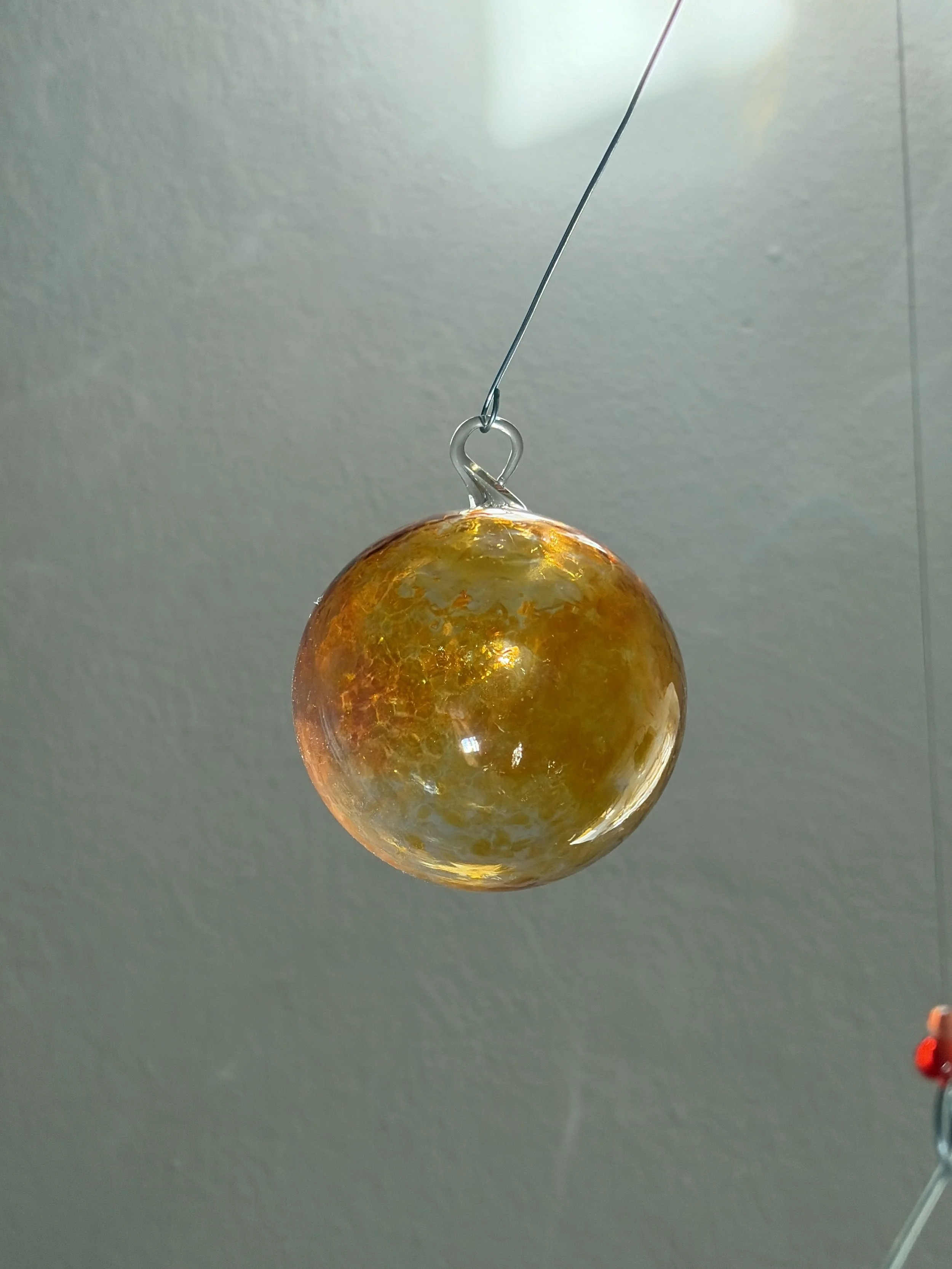 Eine hängende Glasskugel mit goldener und oranger Irisierung, befestigt an einem dünnen Draht vor einer hellgrauen Wand.