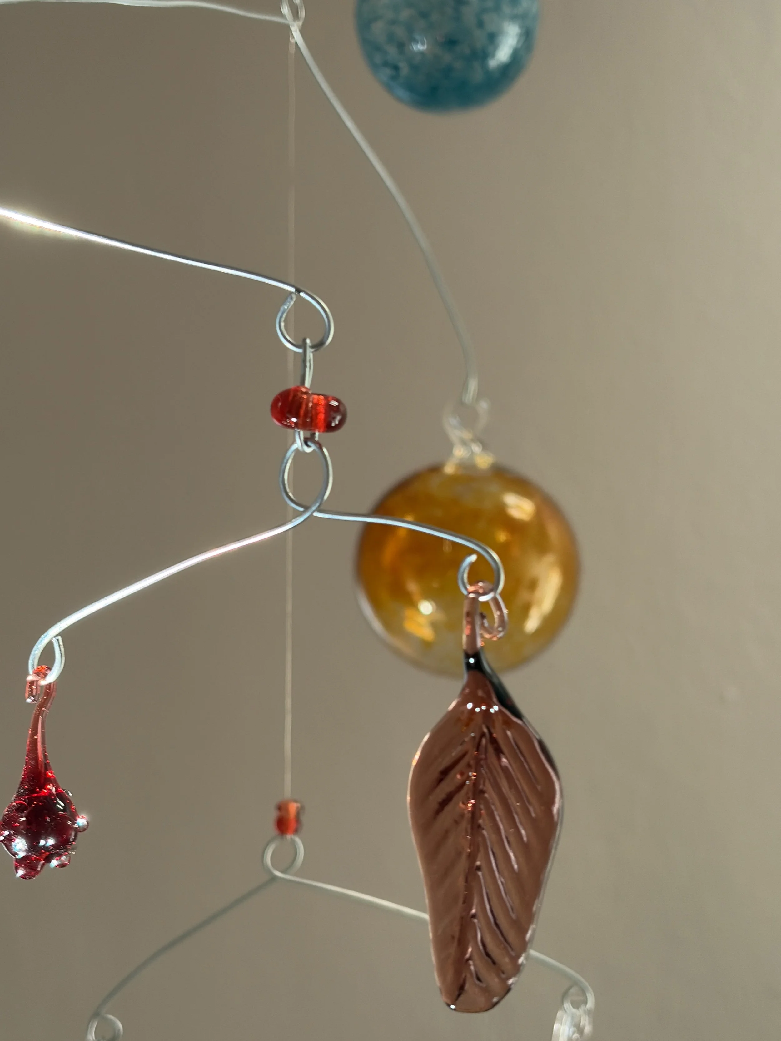 Bunte Glaskunst mit einer sienna brownfarbenen, Blatt-Form, einer roten Glaskugel mit Noppen, einer blauen Kugel und einer bernstein farbenen Kugel, die an Drähten hängen.