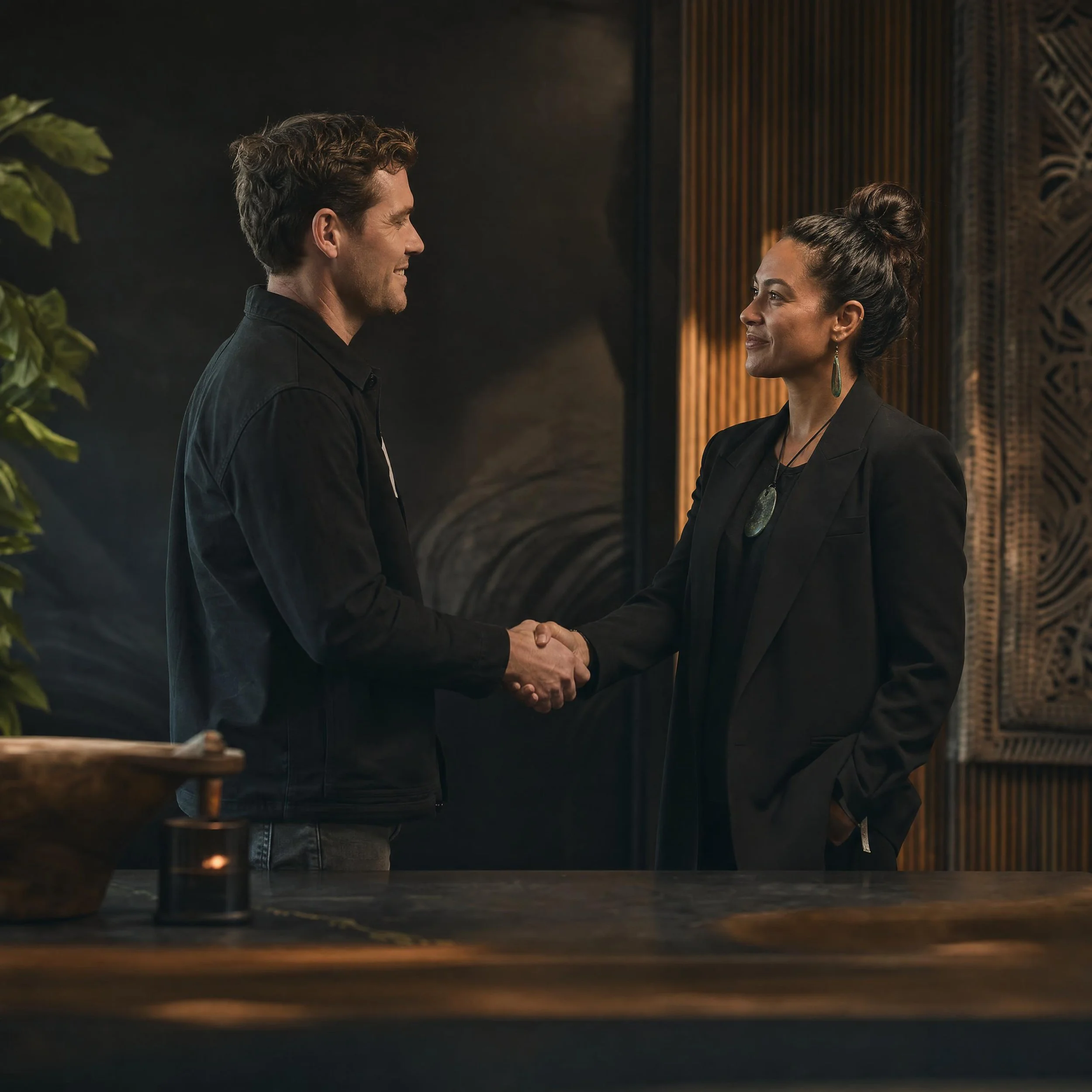 A man and a woman are smiling at each other while shaking hands inside a dimly lit room with dark walls and wooden accents, and green plants are partially visible in the foreground.