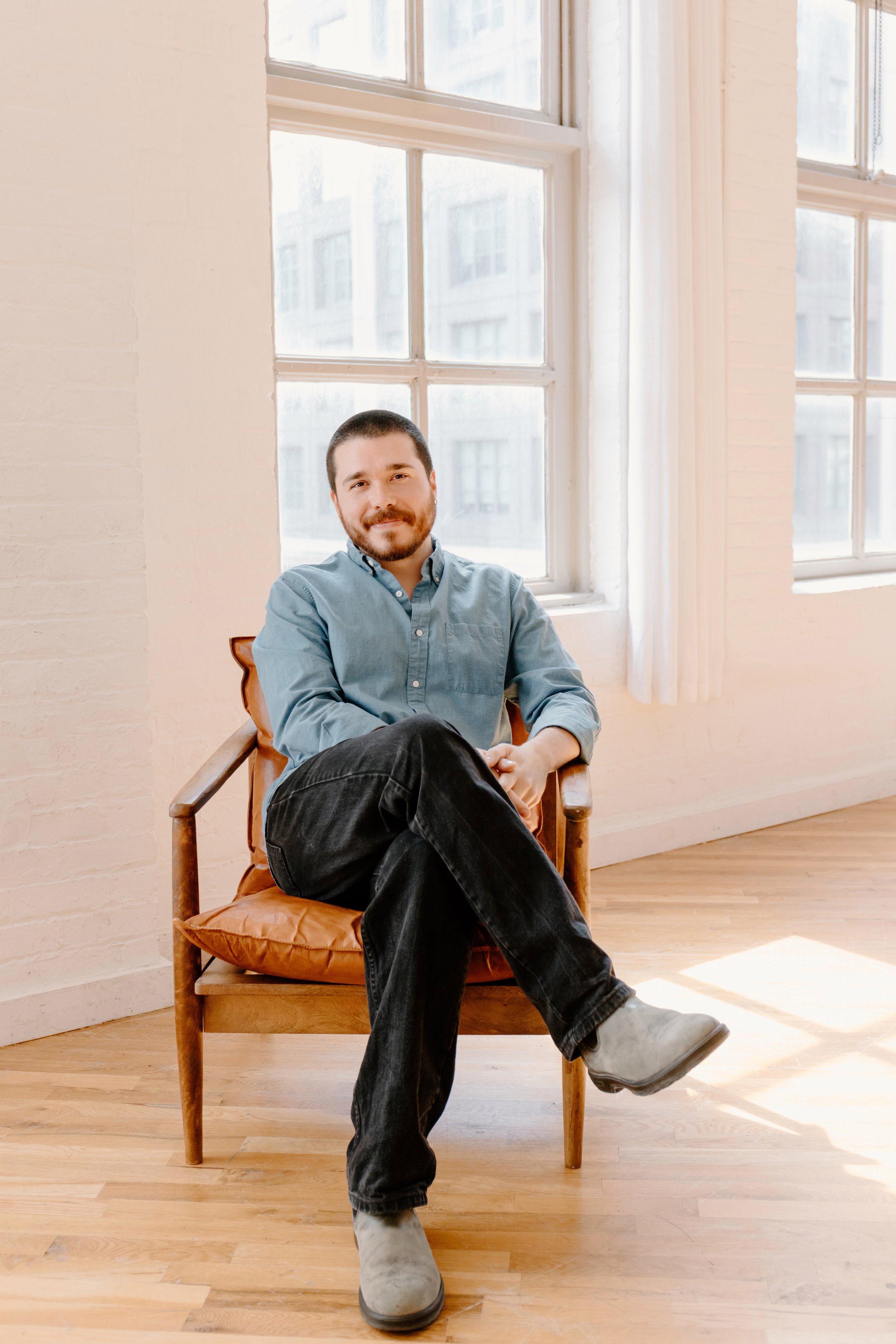 A man with a beard and short hair, wearing a light blue button-up shirt, black jeans, and gray boots, sitting on a wooden chair with a brown cushion in a room with large windows and light-colored walls. He is smiling with his arms crossed.