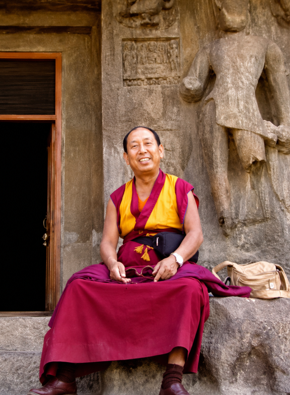 Geshe Sonam Gyaltsen zittend op een steen voor een oude reliëfmuur.