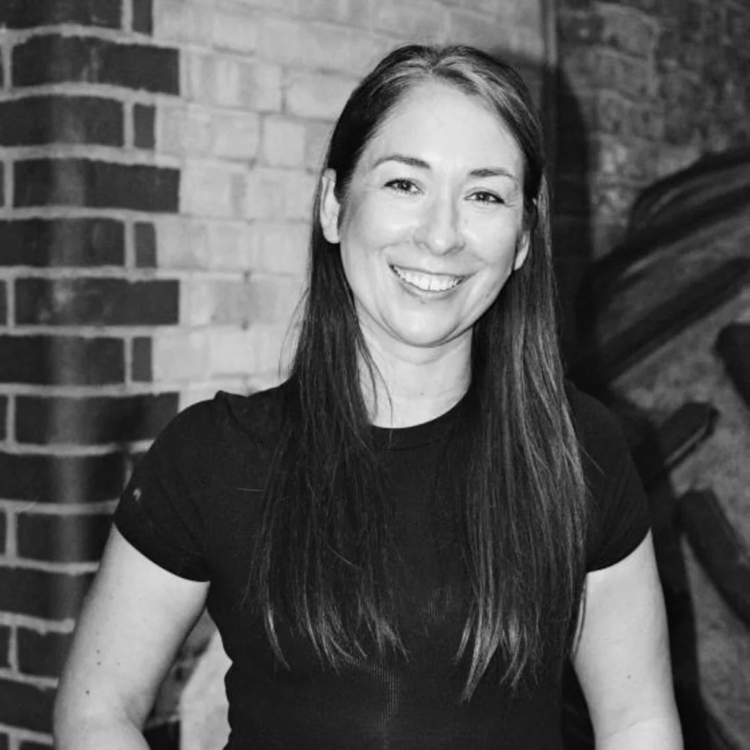 Black and white photo of a smiling woman with long dark hair, wearing a black shirt, standing in front of a brick wall.