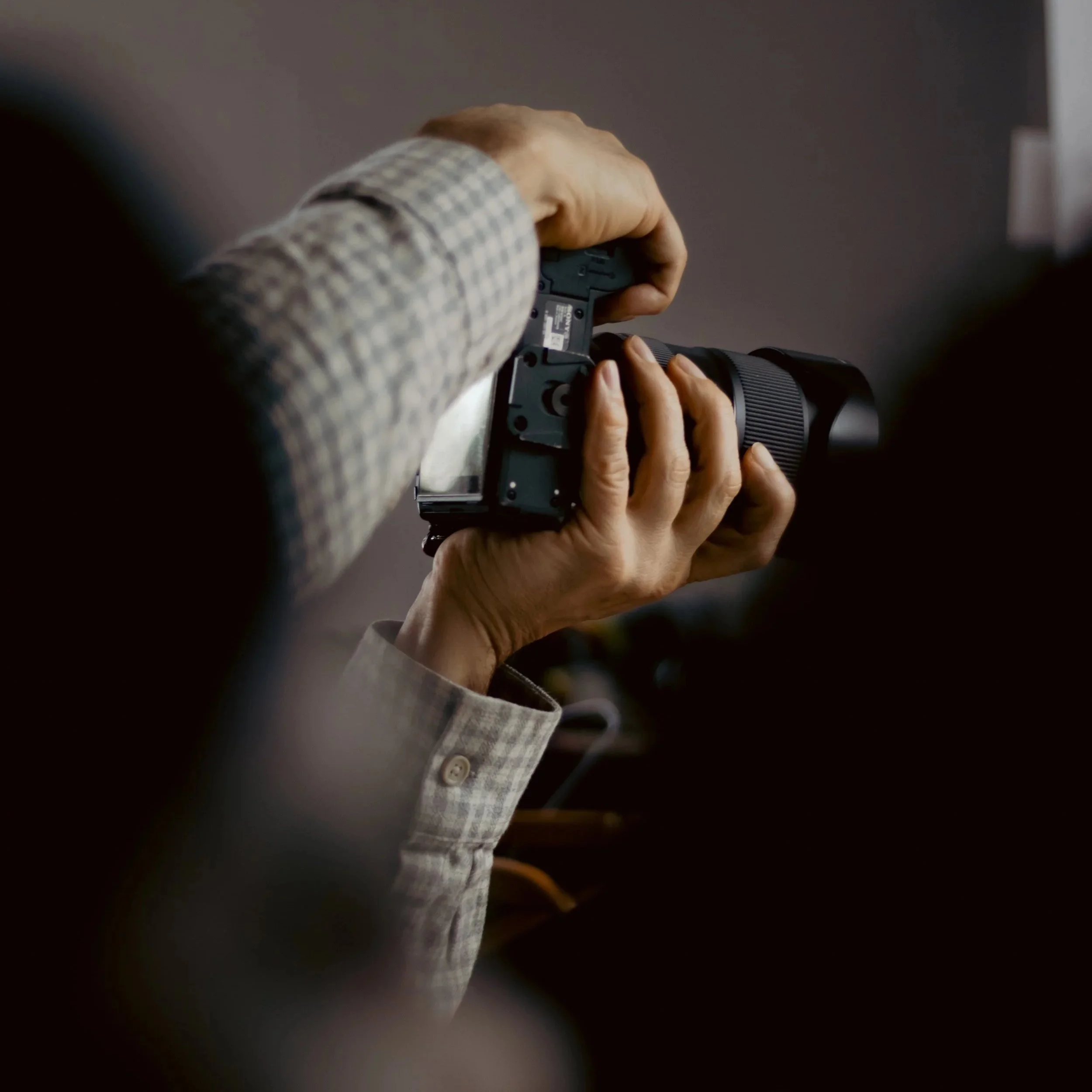A person wearing a gray checkered shirt holding a camera with a large lens, positioned against a dark background, taken at our weekly camera club.