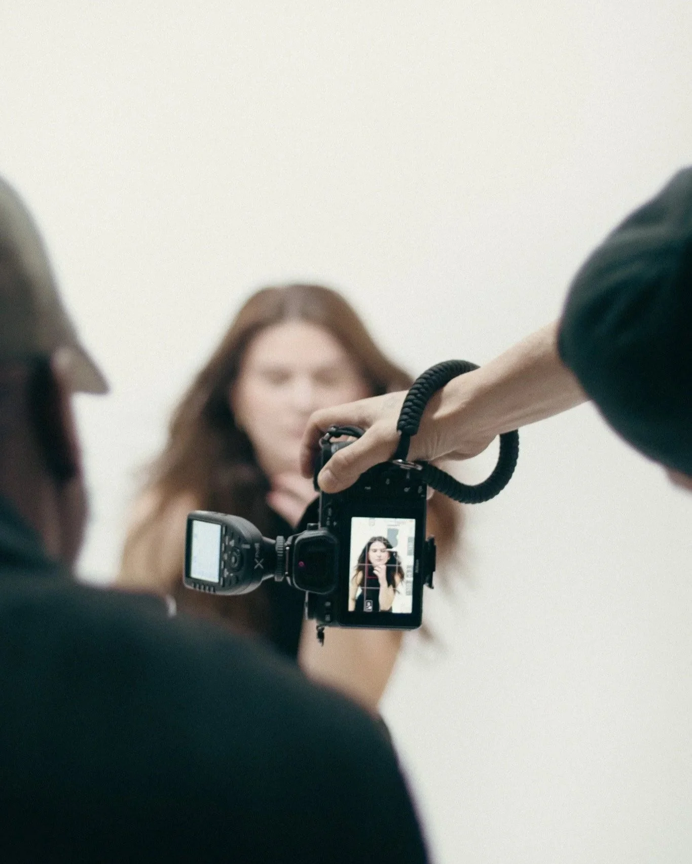 A woman with long brown hair is taking a photo in front of a mirror, with her reflection visible on the camera's screen. Another person's arm is extending from the right, holding the camera steady.