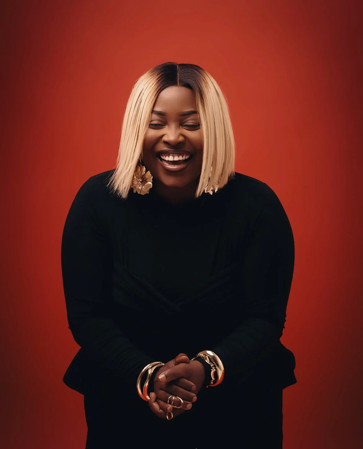 A woman with shoulder-length blonde hair, wearing a black outfit, smiling with closed eyes, standing against a red background, accessorized with gold jewelry and large floral earrings.