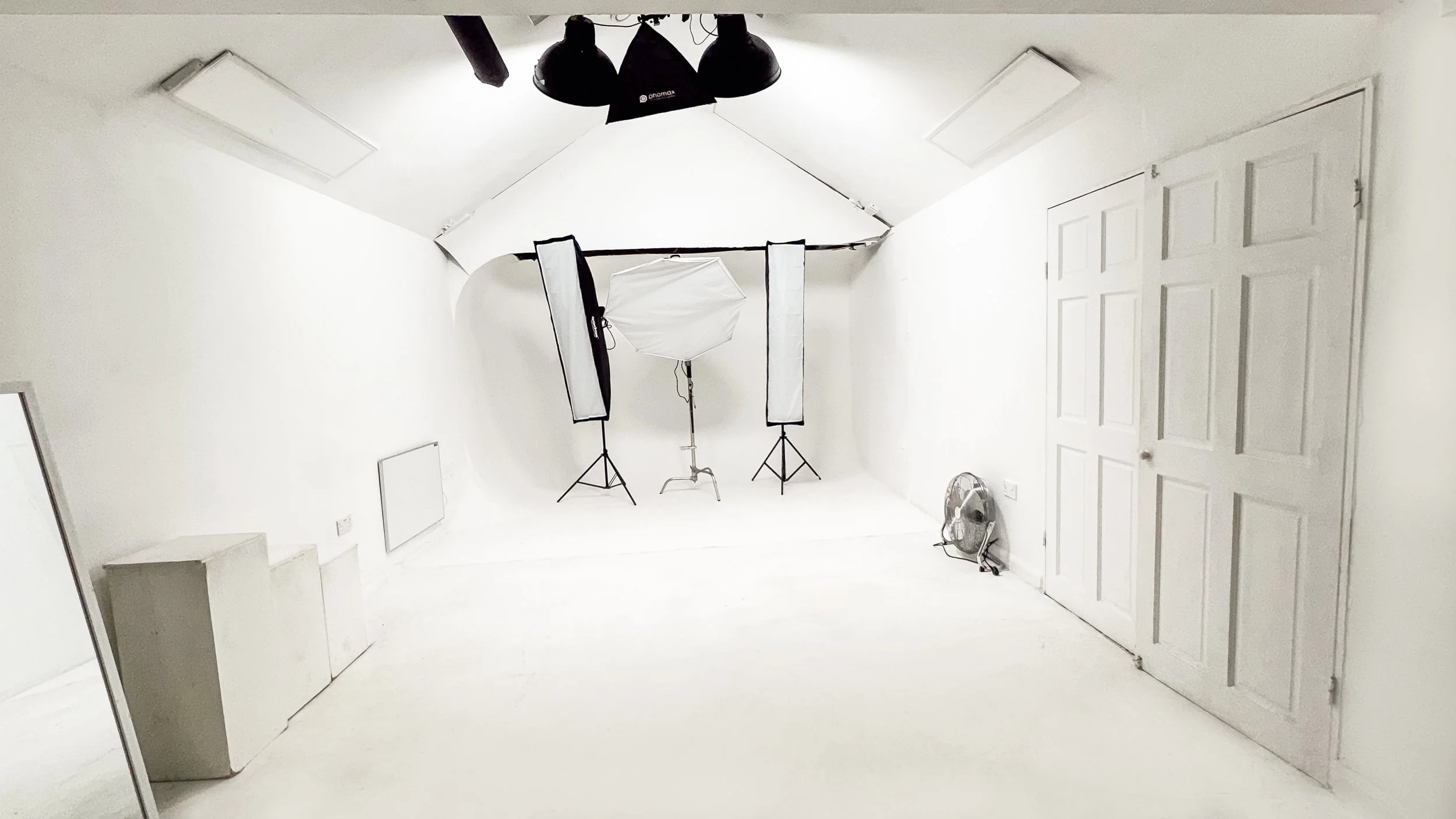 Photography studio with white walls and floor, professional lighting equipment, softbox, and reflectors set up on tripods, a fan on the floor, and a closed door on the right.