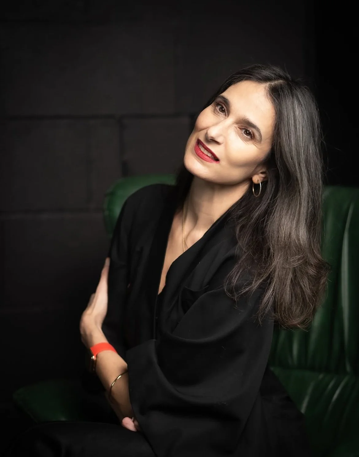 A woman with dark hair and light skin tone, wearing a black outfit, seated against a dark background and green chair, smiling and looking at the camera.