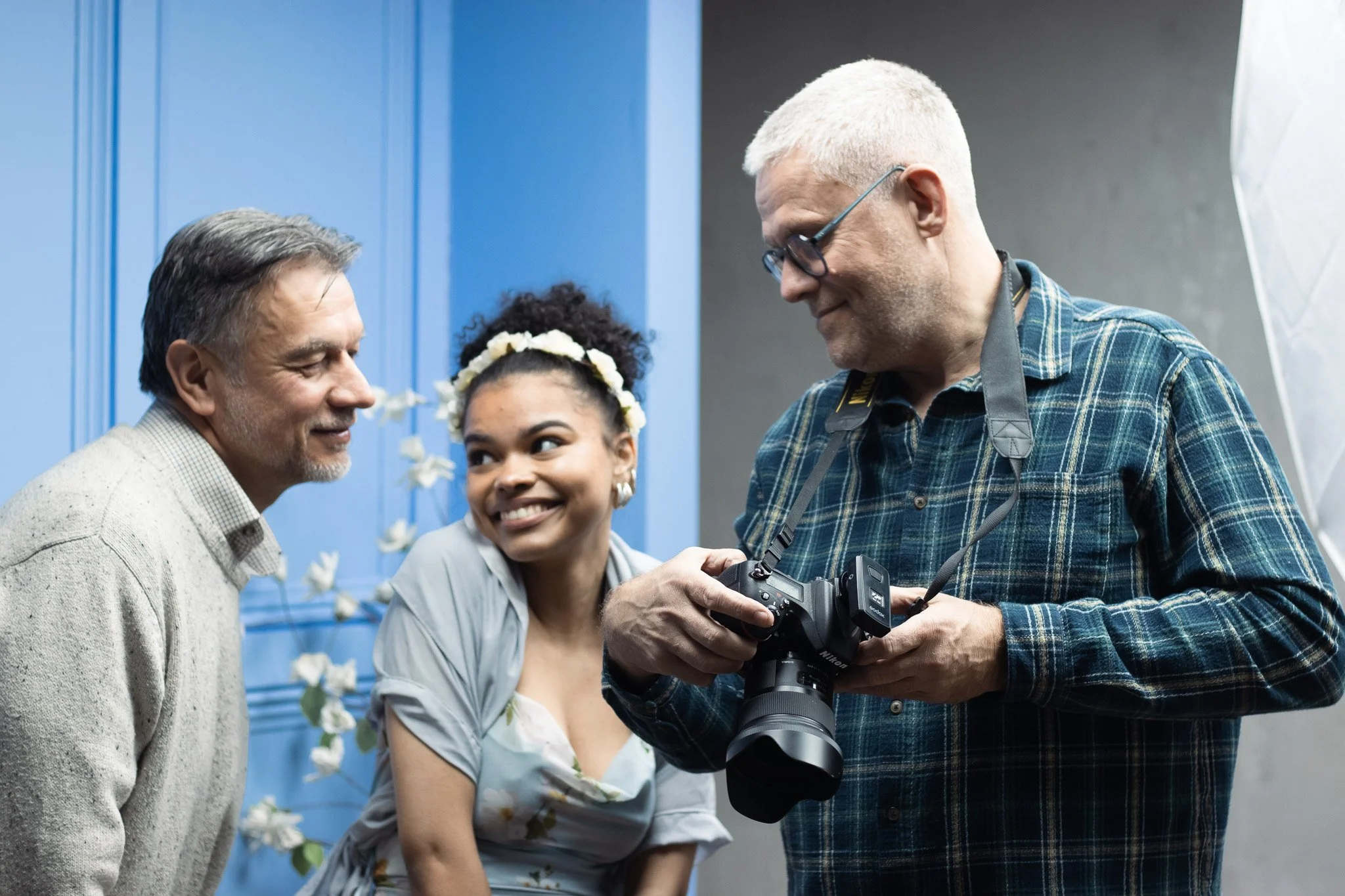 Three people in a photo, two men and one woman, standing together indoors. The woman is smiling and wearing a flower crown, while the man on the right is holding a camera and looking down at it. The man on the left is looking at the woman, all engaged in a friendly interaction.