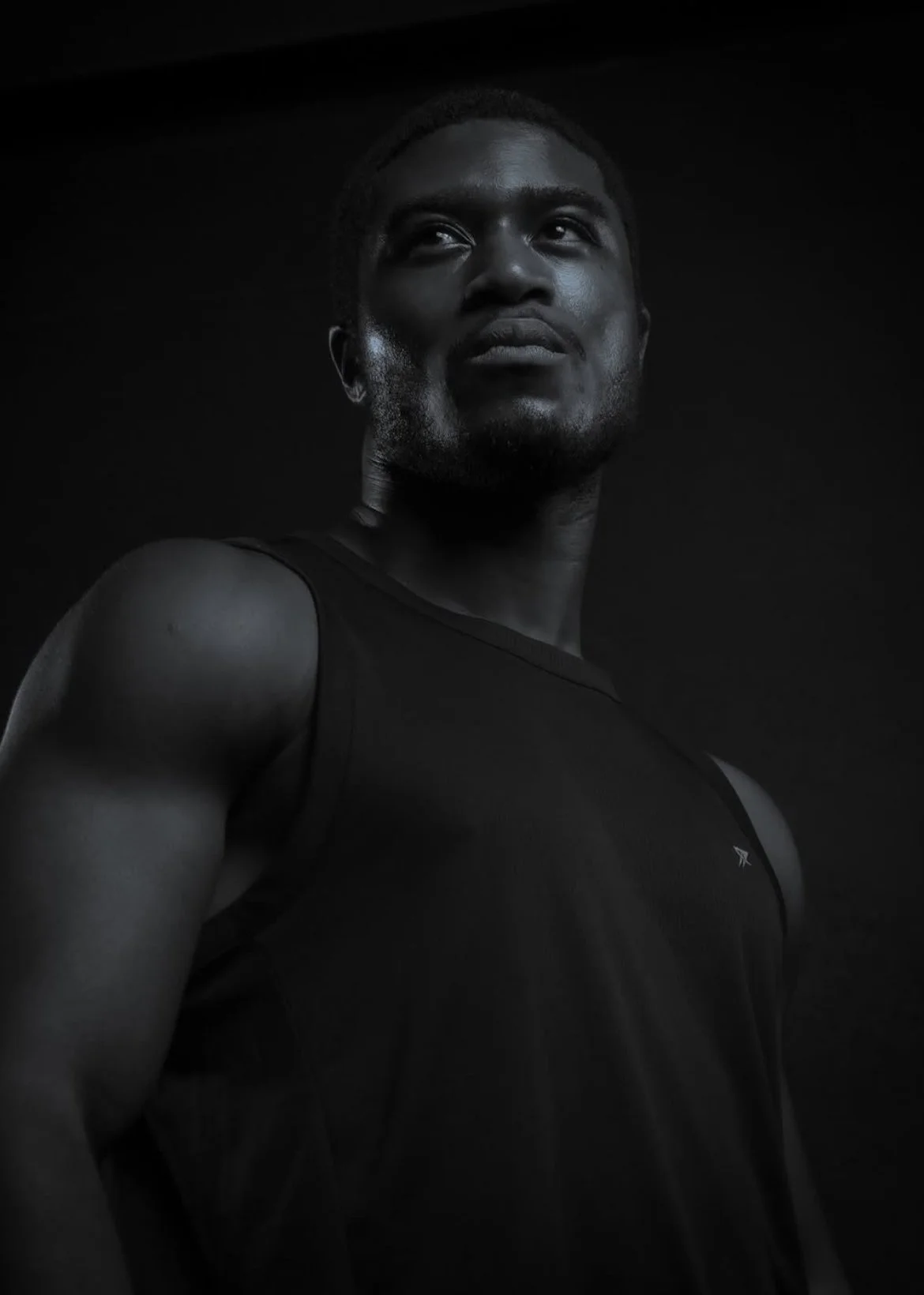 Black man wearing athletic tank top looking upward against a dark background.