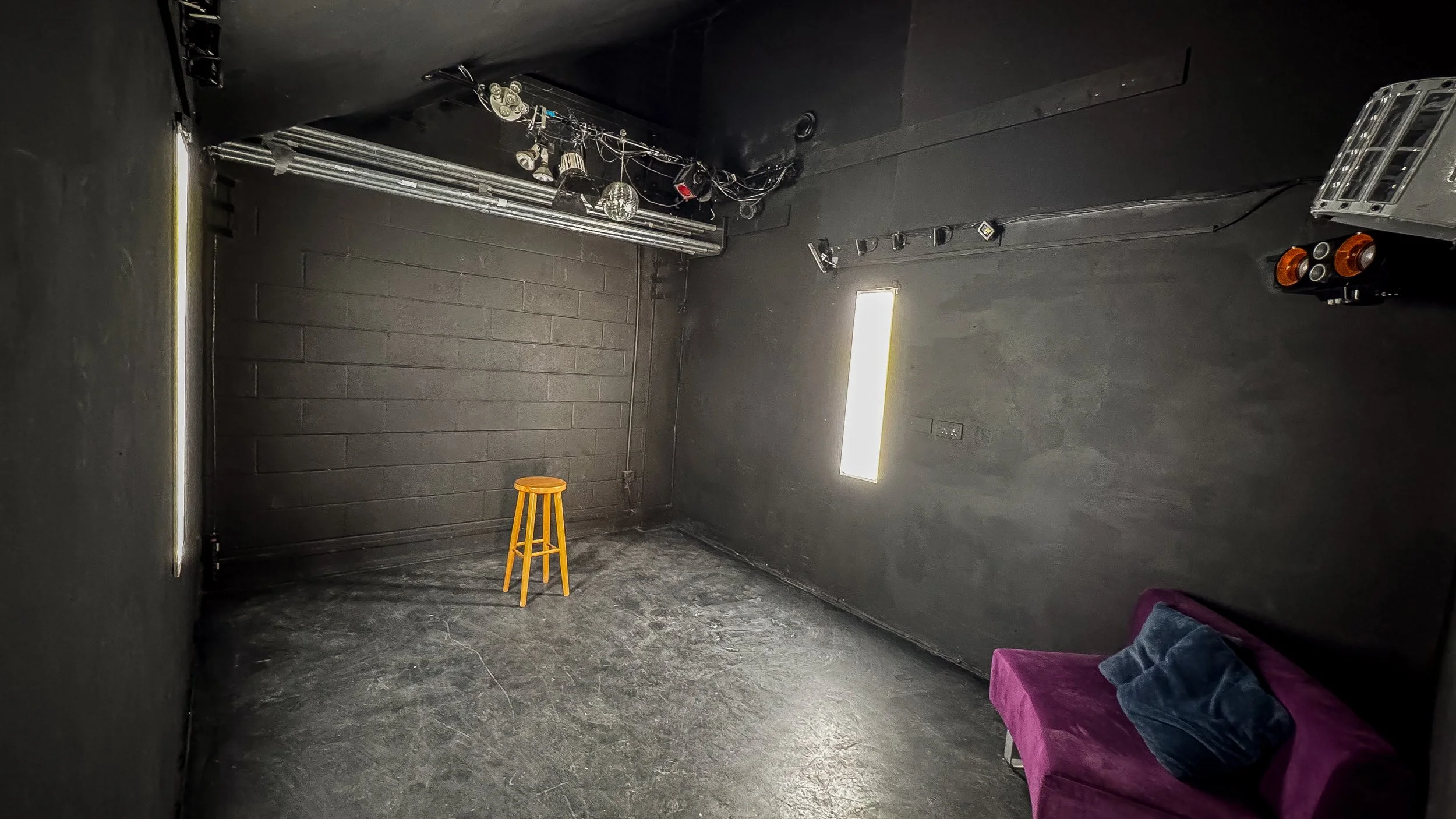 Empty indoor space with black walls and floor, a lone yellow stool, purple sofa with pillows, and stage lighting equipment hanging from ceiling.