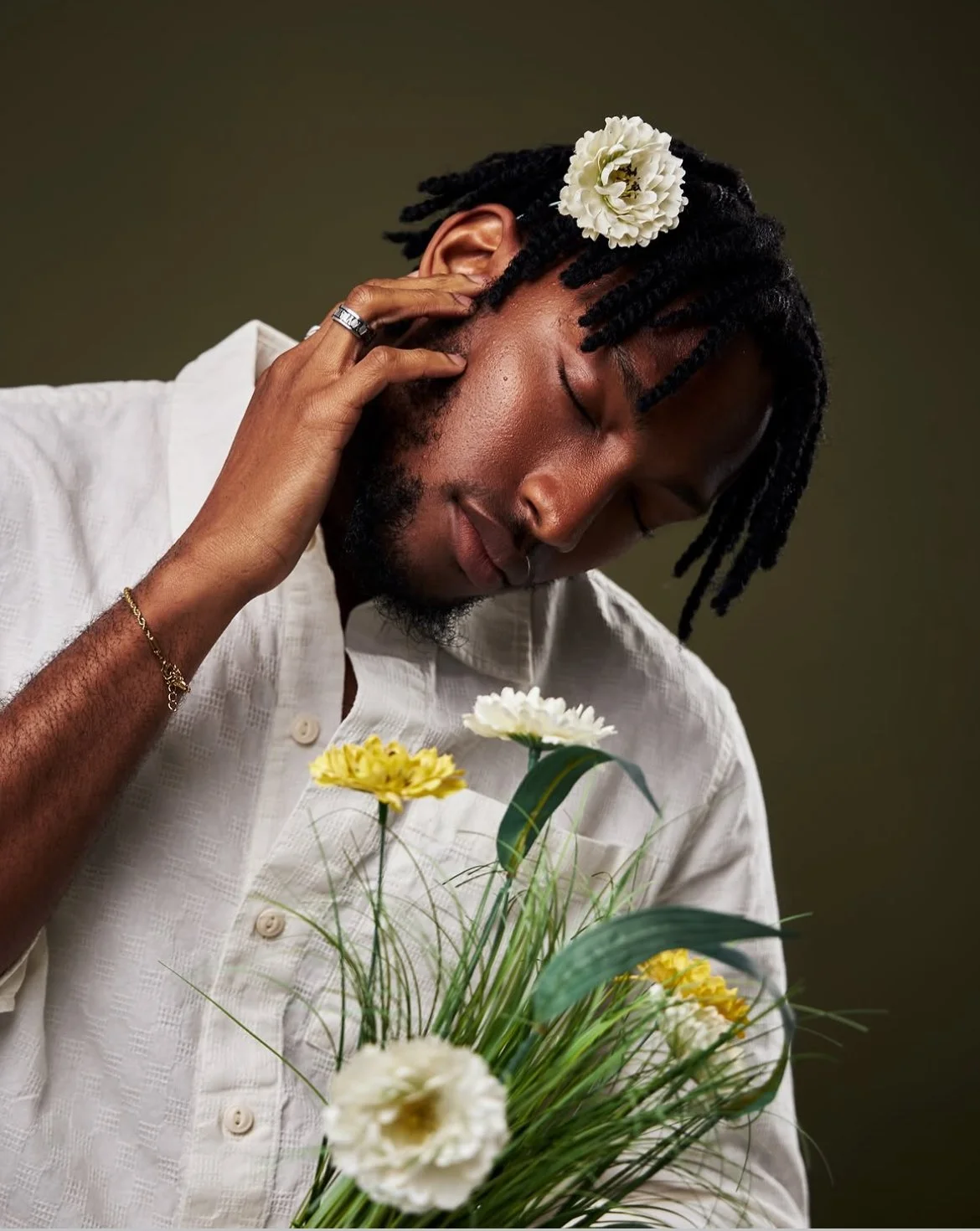 A man with closed eyes, wearing a white shirt, touching the side of his face with one hand, has black dreadlocks, a white flower in his hair, and is holding a bouquet of yellow and white flowers.