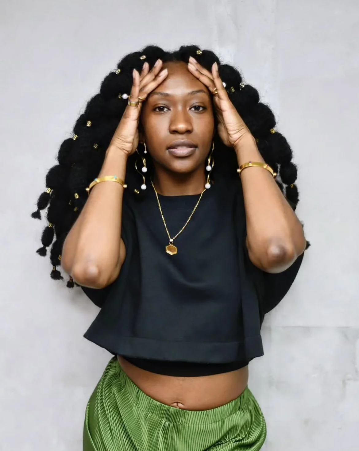 Woman with voluminous black hair styled in large twists adorned with gold and white hair accessories, wearing gold jewelry, a black crop top, and green patterned pants, posing with hands on her head against a plain background.