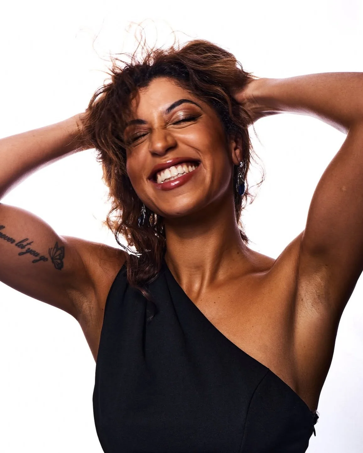 Woman with short wavy brown hair, smiling with eyes closed, holding her hands behind her head, wearing earrings and a black one-shoulder top, standing against a white background.