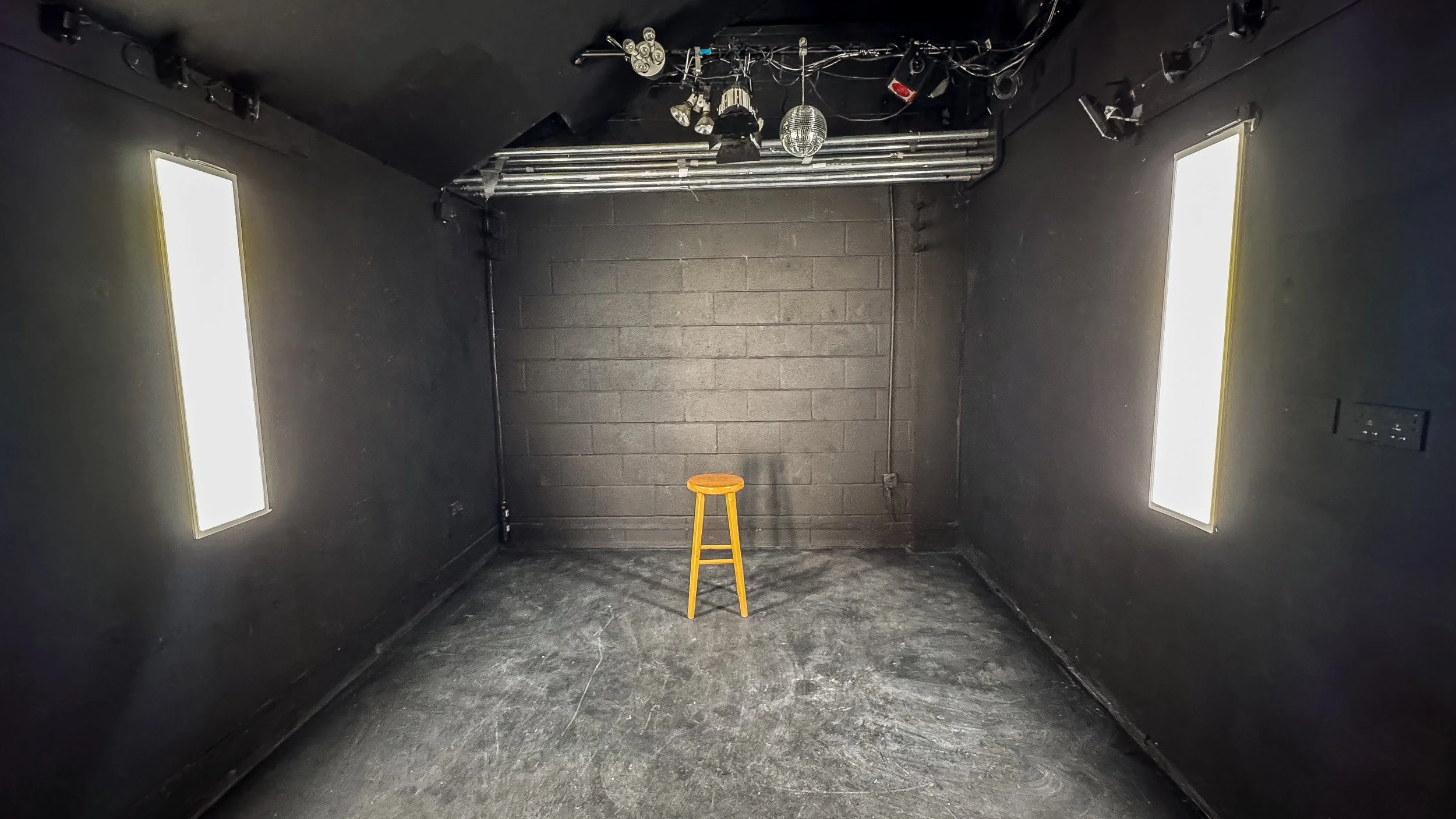 An empty black room with two vertical bright light panels on the left and right walls, a yellow stool in the center, and various stage lighting equipment hanging from the ceiling.