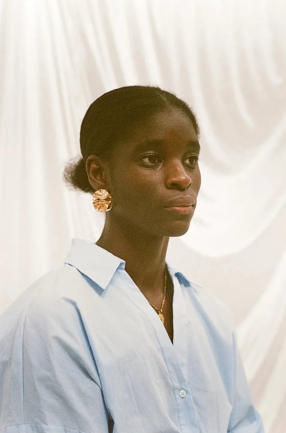 Portrait of a Black woman with short hair, wearing a light blue shirt, large gold earrings, and a gold necklace, against a white background.