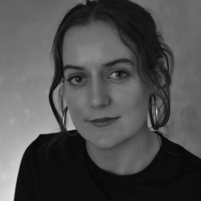 Black and white portrait of a young woman with wavy hair, wearing hoop earrings and a dark top, looking at the camera.