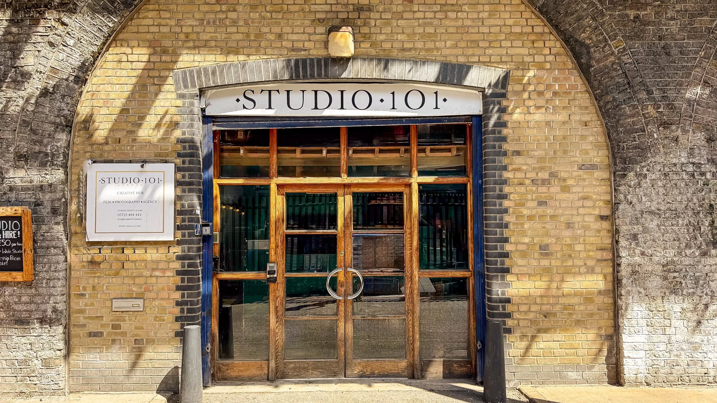 Entrance of a brick building with a sign that reads 'STUDIO 101' above the door. Wooden framed glass door with metal handles, and a smaller bulletin board to the left with more information about the studio.