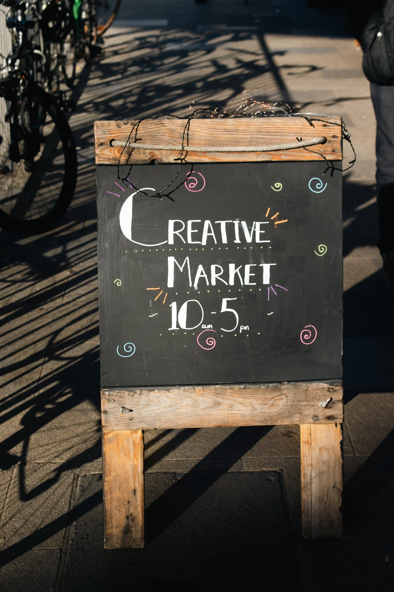A chalkboard sign with colorful drawings and text advertising a creative market from 10 am to 5 pm, mounted on a wooden stand outdoors on a sunny day.