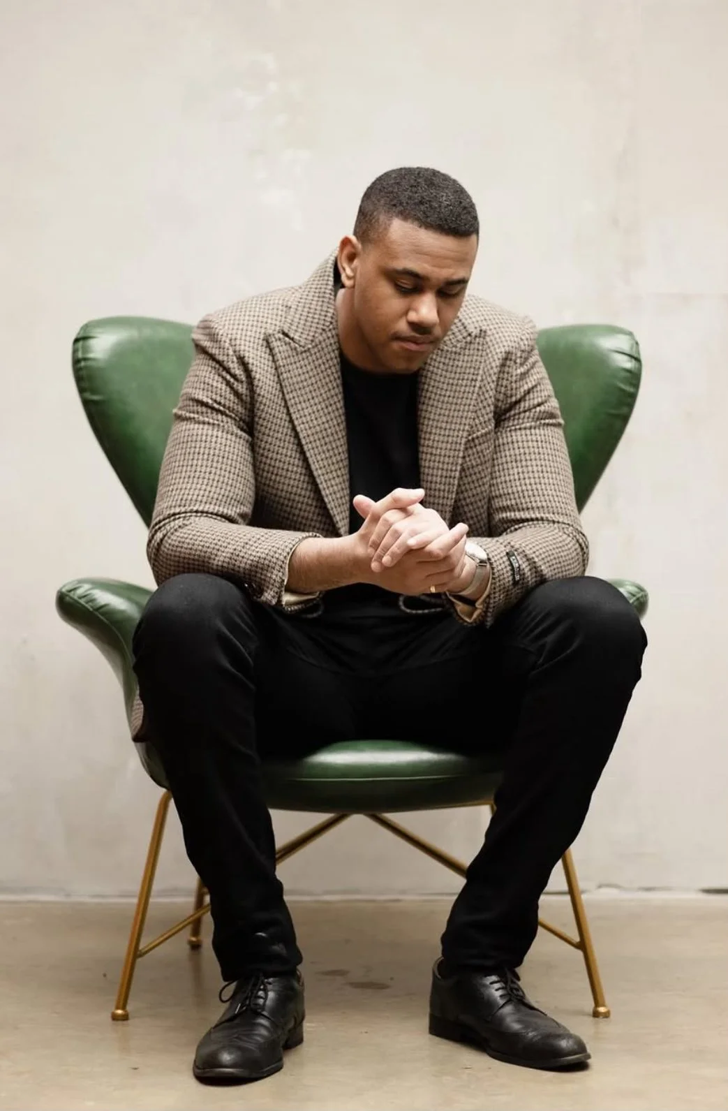 A man sitting in a green chair, wearing a checked blazer and black pants, with his hands clasped and head bowed.