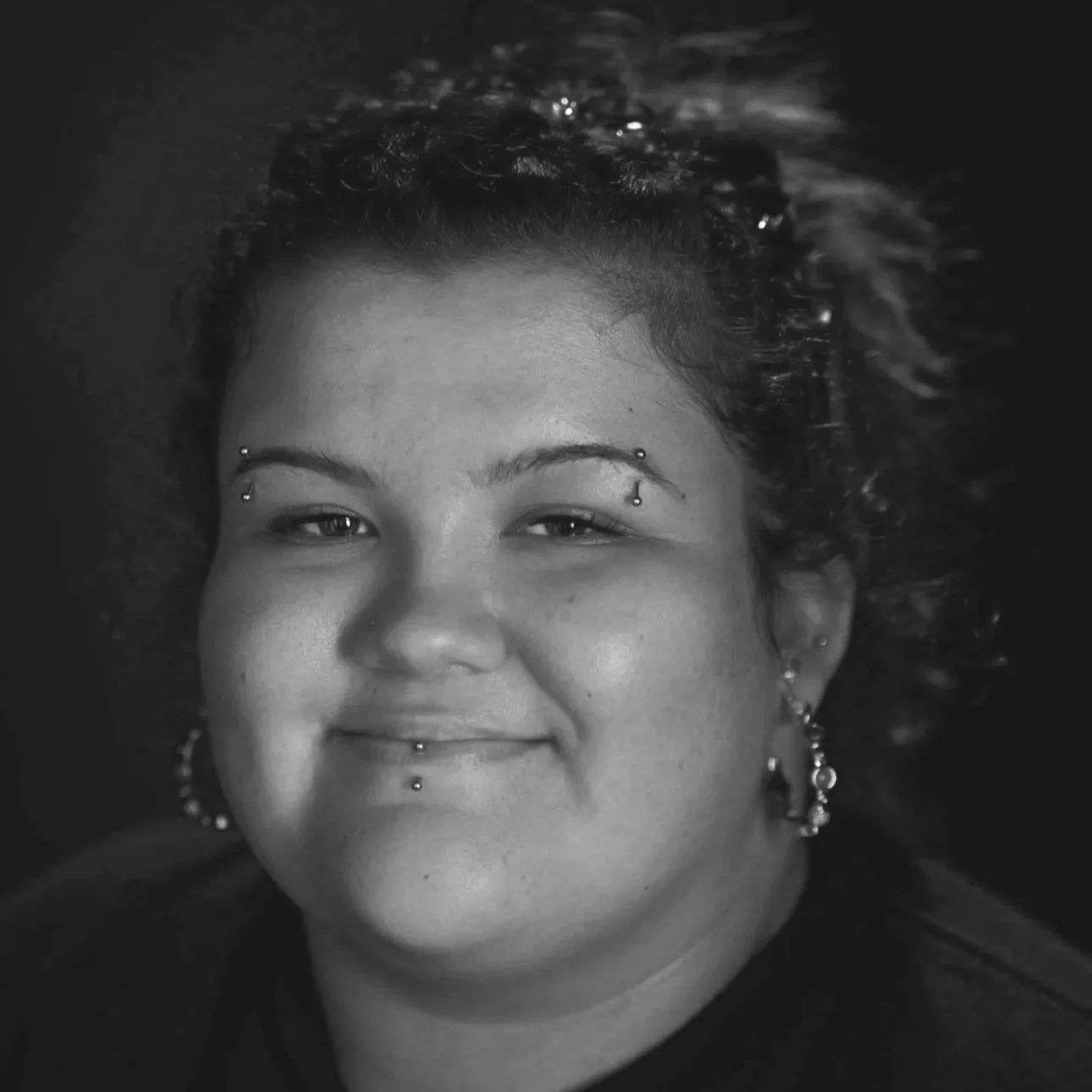 Black and white portrait of a smiling woman with multiple facial piercings and earrings, with curly hair.