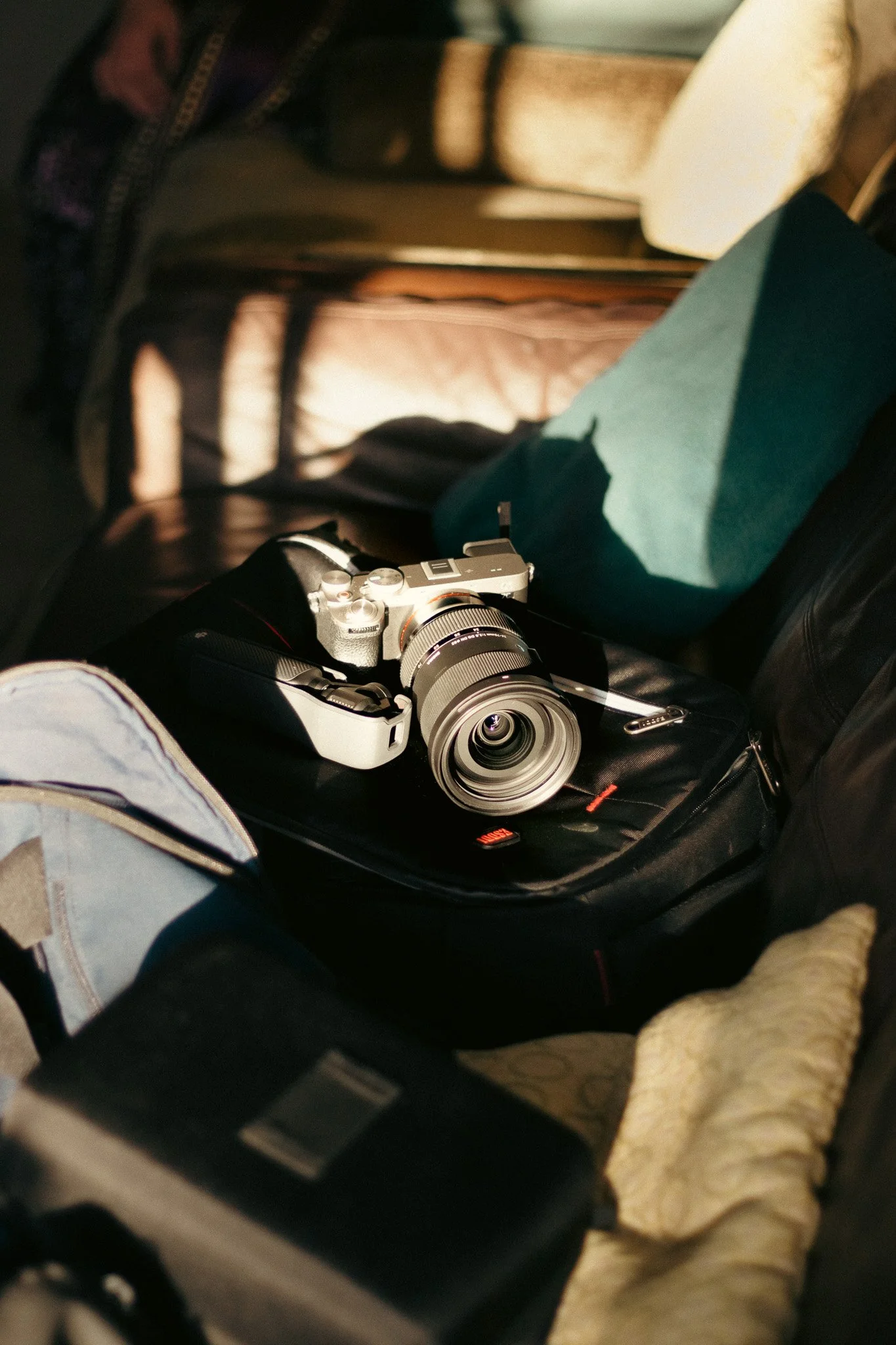 A digital camera with silver and black body and a large lens, resting on a black camera bag inside a vehicle with a tan seat and a teal-colored object nearby.