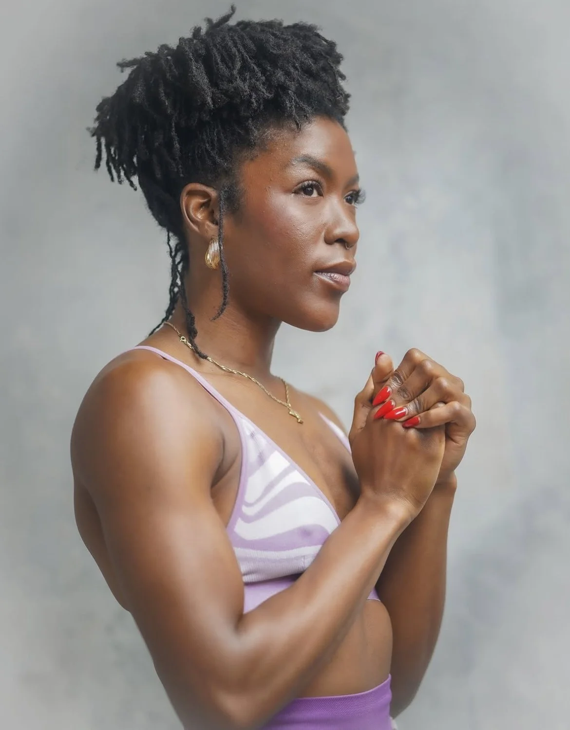 A woman with dark skin and natural hair styled in dreadlocks, wearing a pink and white sports bra, gold jewelry, and red nail polish, standing against a neutral background with her hands clasped in front of her chest.