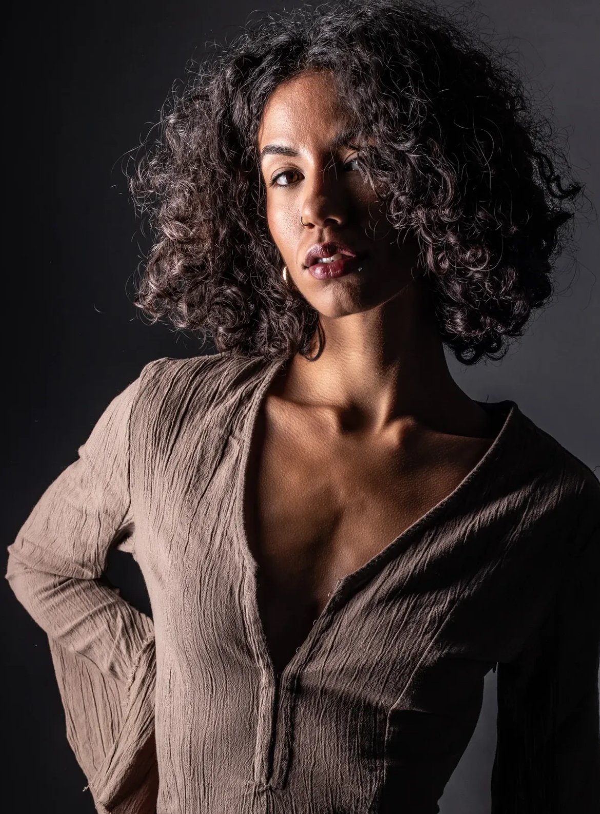 A woman with curly hair and a nose piercing wearing a brown, textured shirt against a dark background.