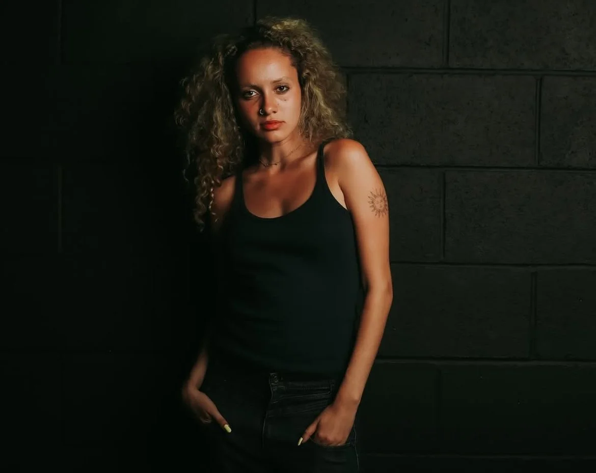 Young woman with curly hair, wearing a black tank top and jeans, standing against a dark brick wall, looking at the camera.