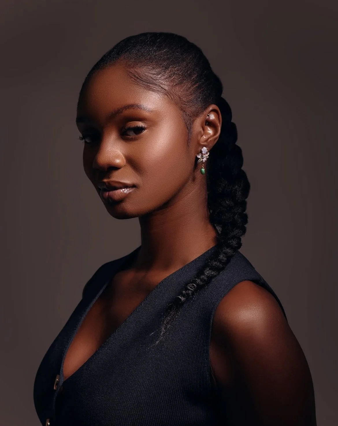 Portrait of a young woman with dark hair styled in a braid, wearing earrings, and a sleeveless dark top against a plain dark background.